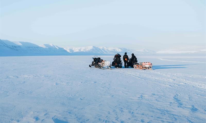 Midt i et hvitt og snødekt landskap, står tre snøscootere. ene personen står mellom de to andre som fortsatt sitter på snøscooteren.