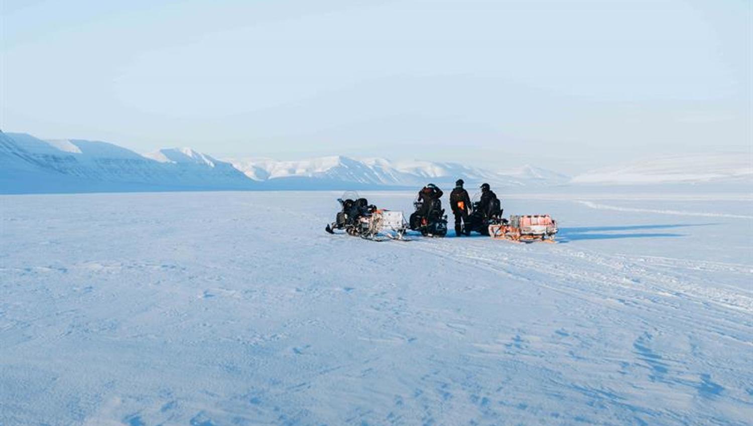Midt i et hvitt og snødekt landskap, står tre snøscootere. ene personen står mellom de to andre som fortsatt sitter på snøscooteren.