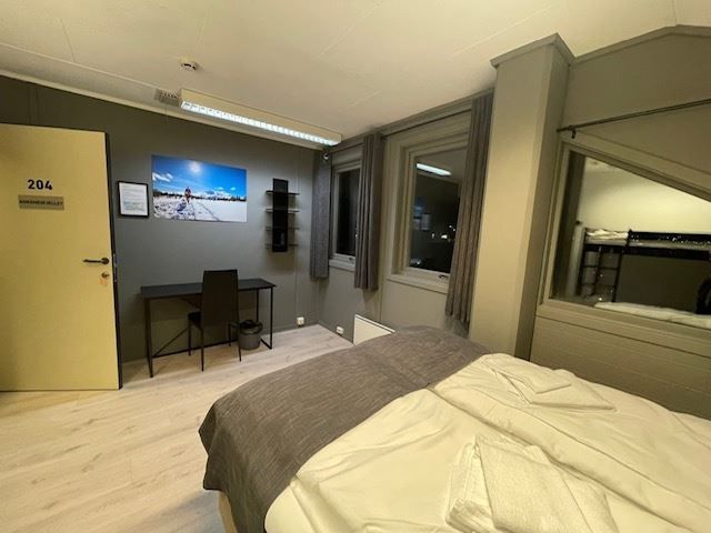 Simple hotel-style bedroom with a made bed, desk and chair, wall art, and windows with curtains at night.