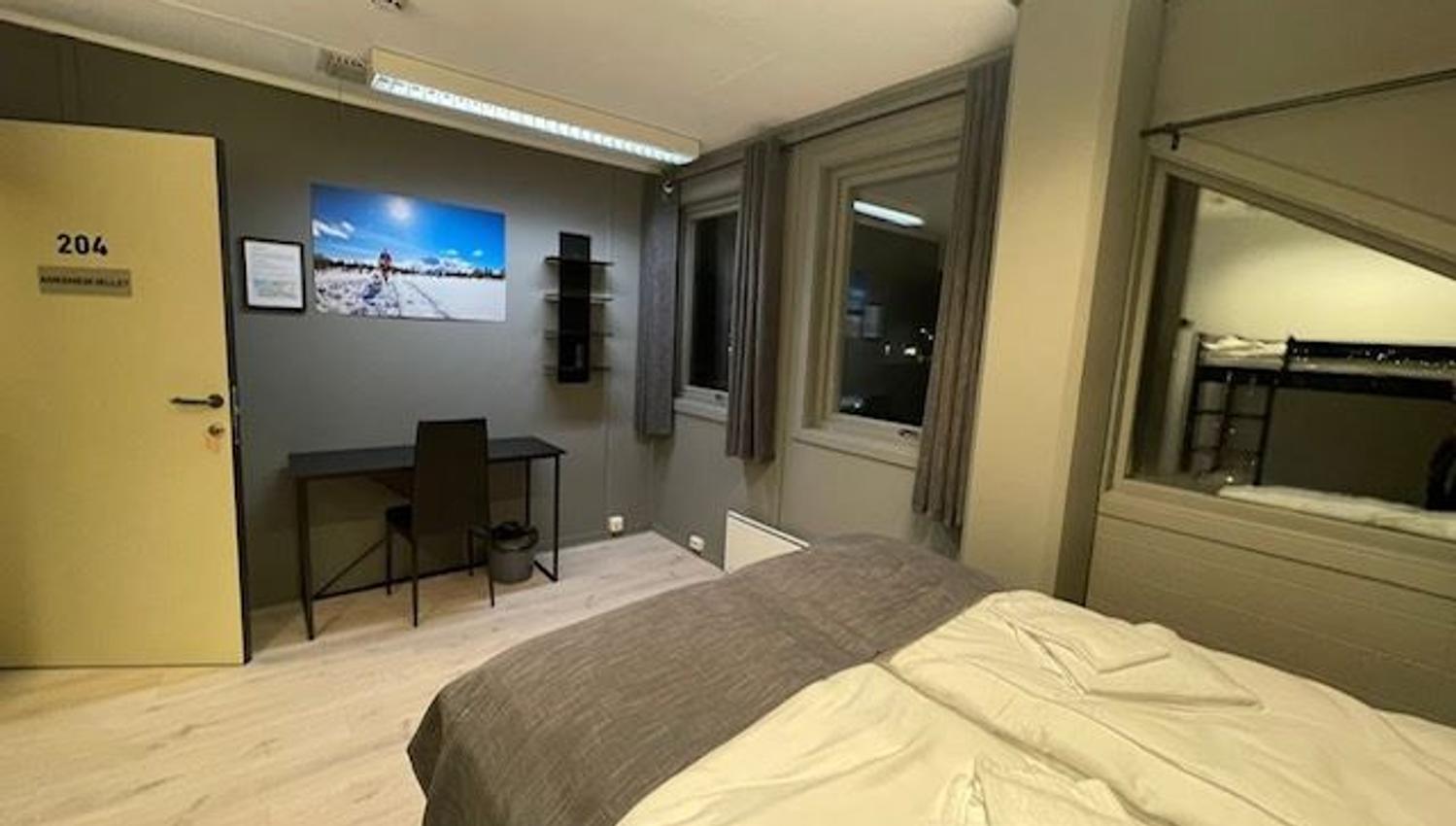 Simple hotel-style bedroom with a made bed, desk and chair, wall art, and windows with curtains at night.