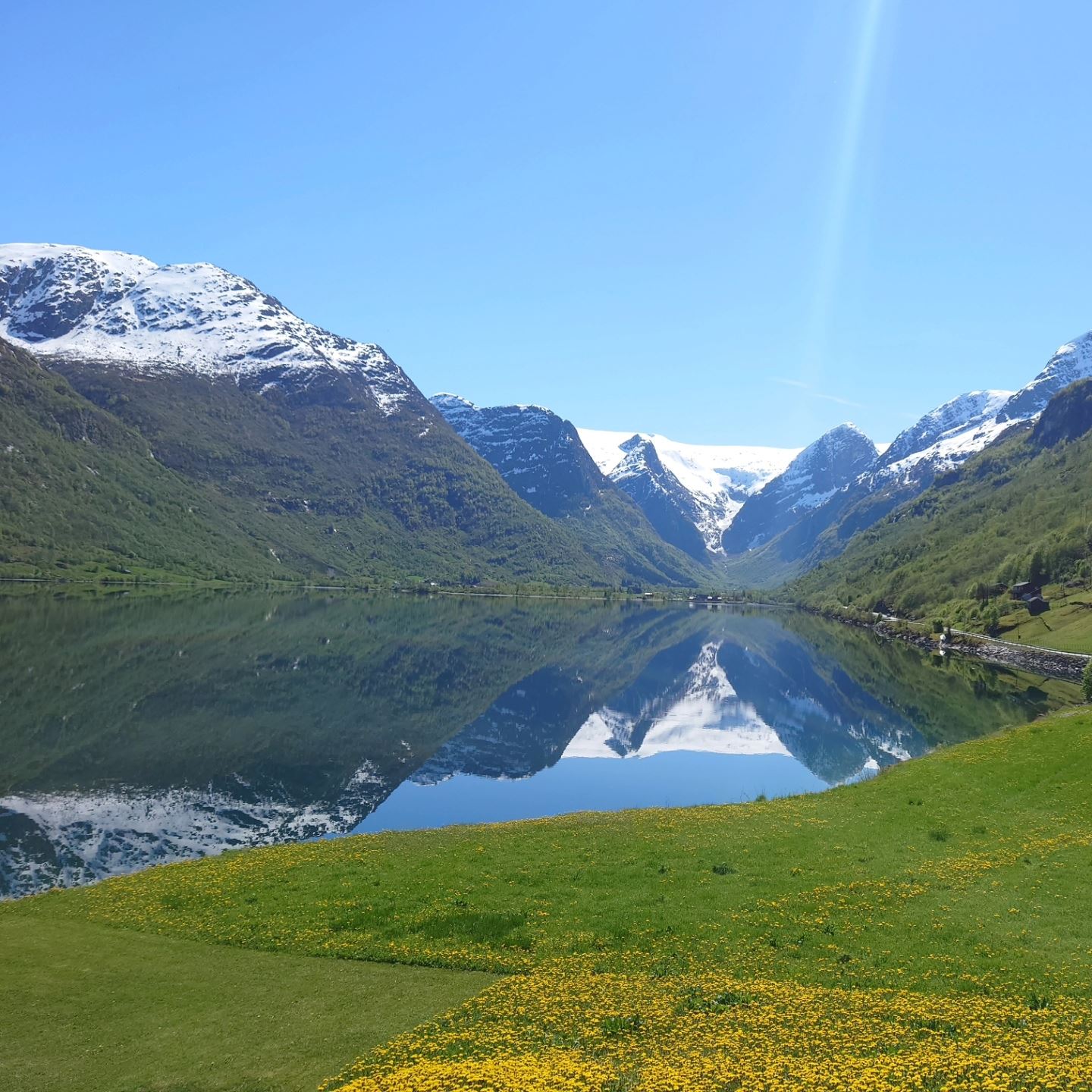 Snow-capped mountains and green slopes mirrored in a calm fjord, with a grassy meadow and blue sky at Aabrekk Gard Trollbu in Oldedalen valley..