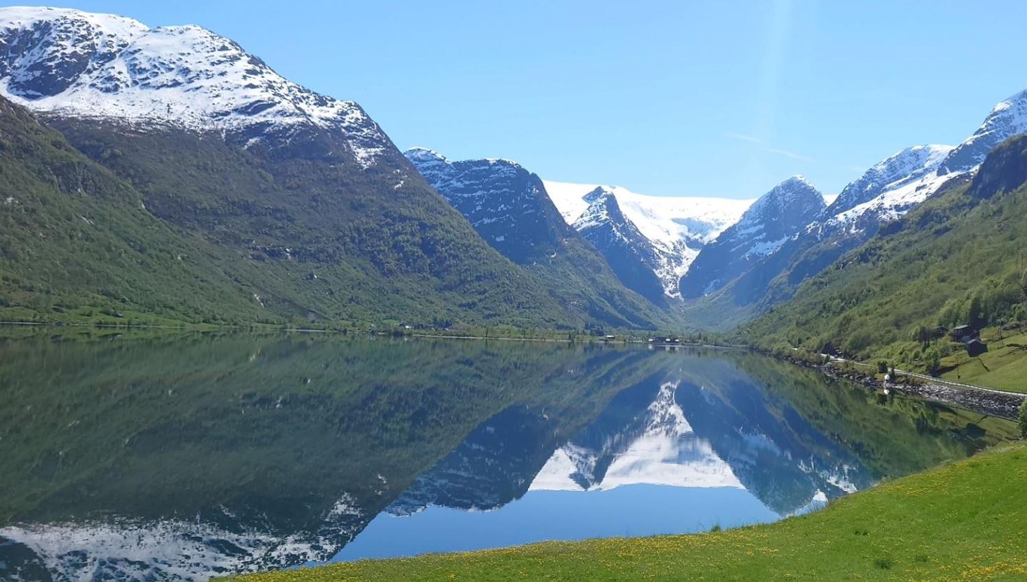Snow-capped mountains and green slopes mirrored in a calm fjord, with a grassy meadow and blue sky at Aabrekk Gard Trollbu in Oldedalen valley..
