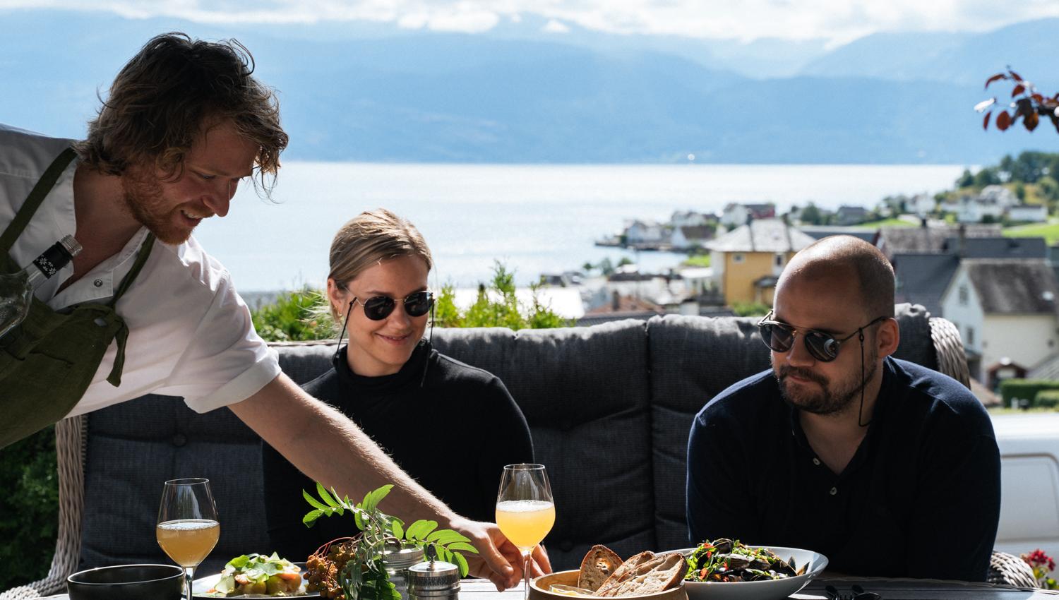 Host serving local food outdoors with fjord view at Eldhuset Farm Restaurant.