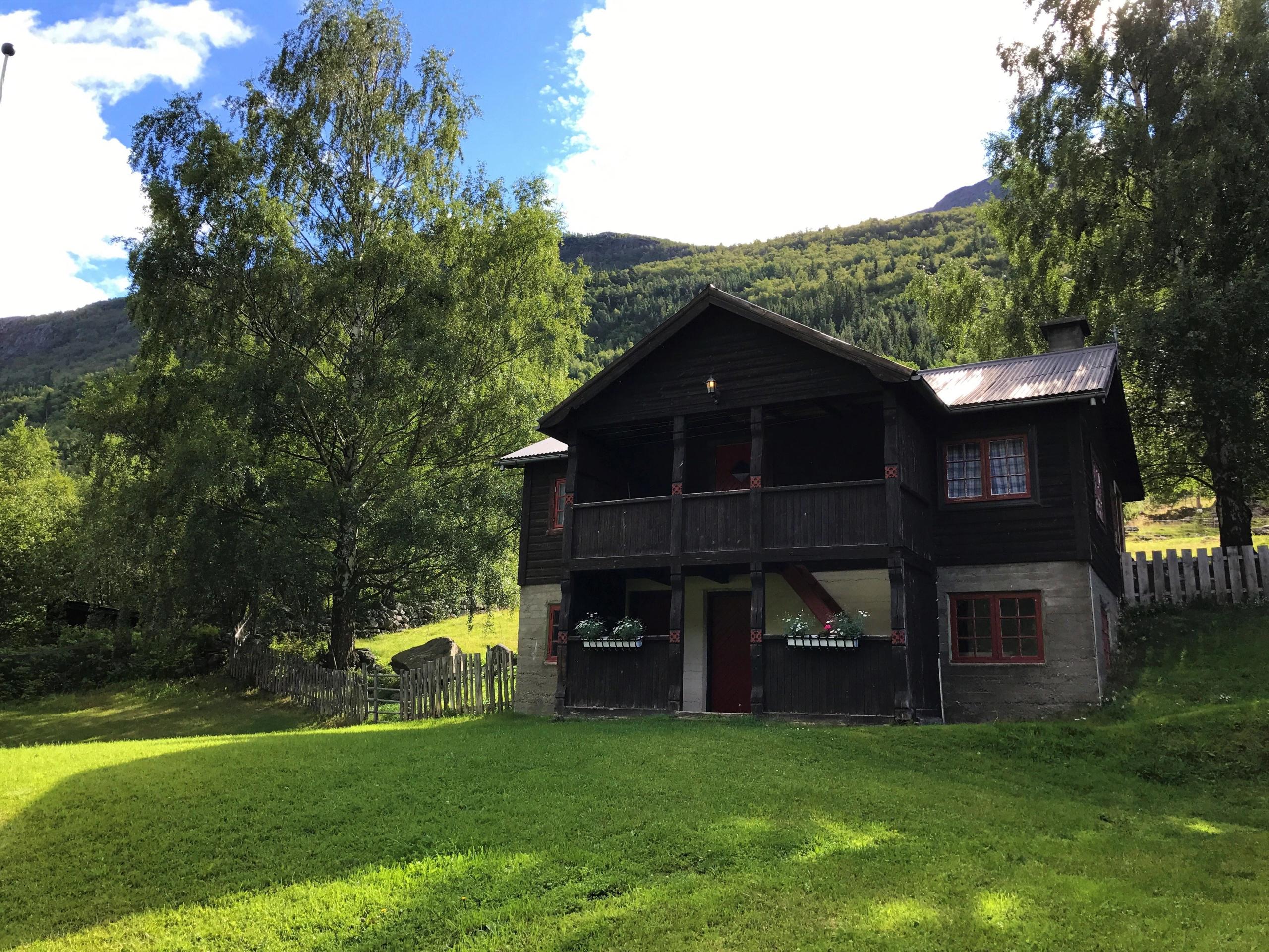 Dark wooden cabin with balcony on a green lawn, framed by leafy trees and sunlit mountains under blue sky..