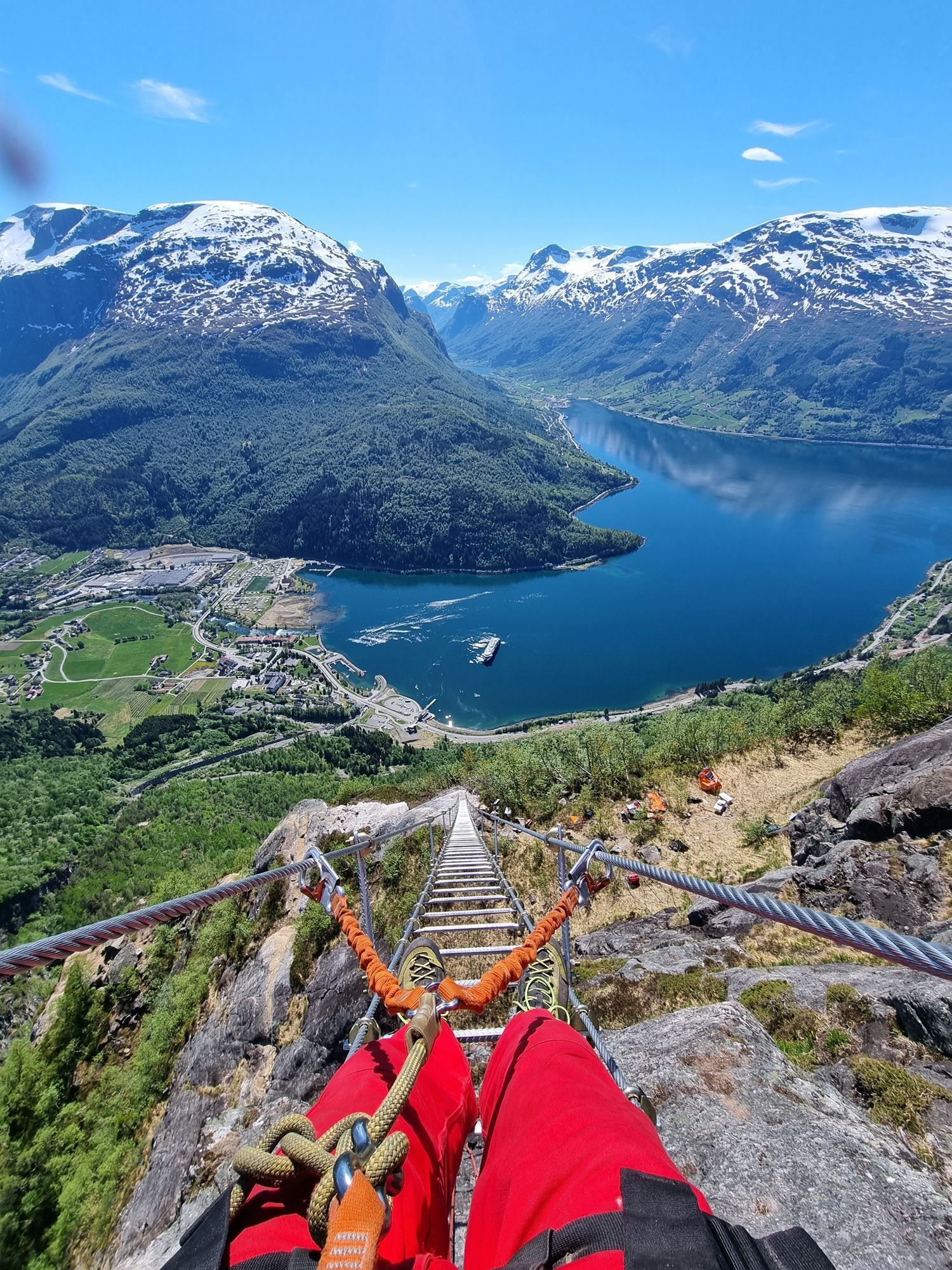 View down a steep metal ladder with safety cables toward a blue fjord, green valley, and snowcapped mountains..