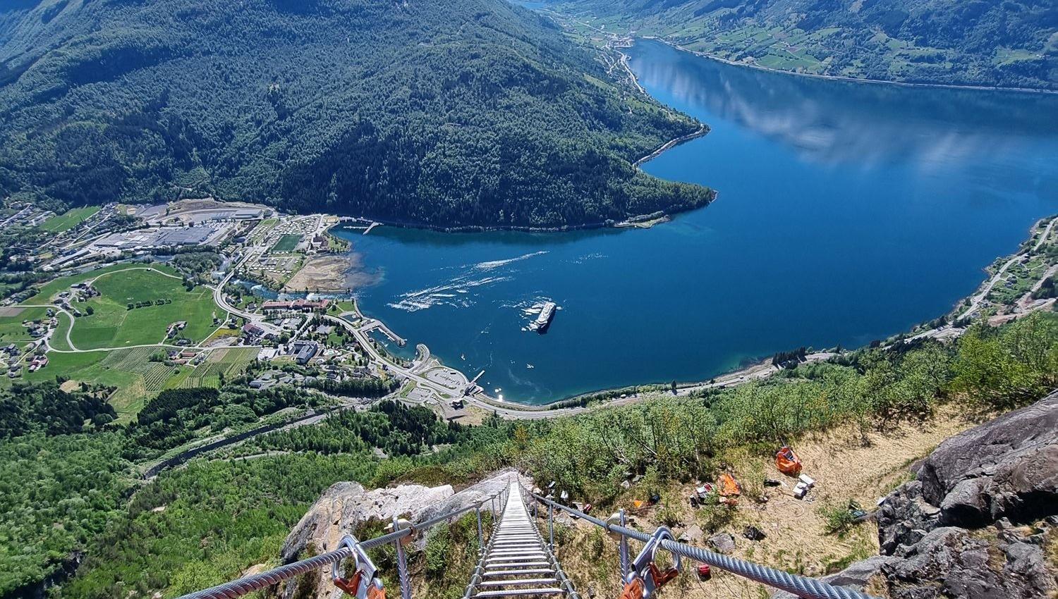 View down a steep metal ladder with safety cables toward a blue fjord, green valley, and snowcapped mountains..