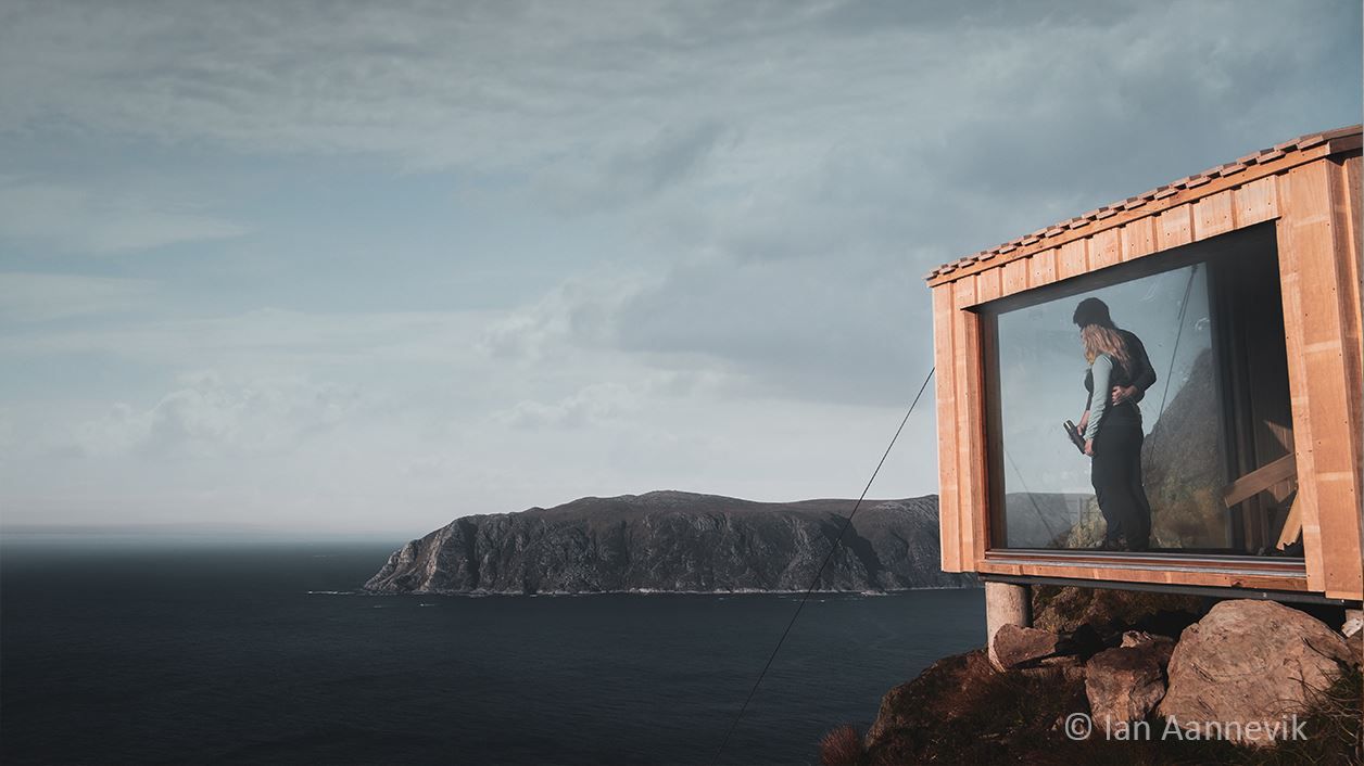 A couple stands in a large window of a wooden cabin on a rocky cliff, overlooking the sea and distant hills..