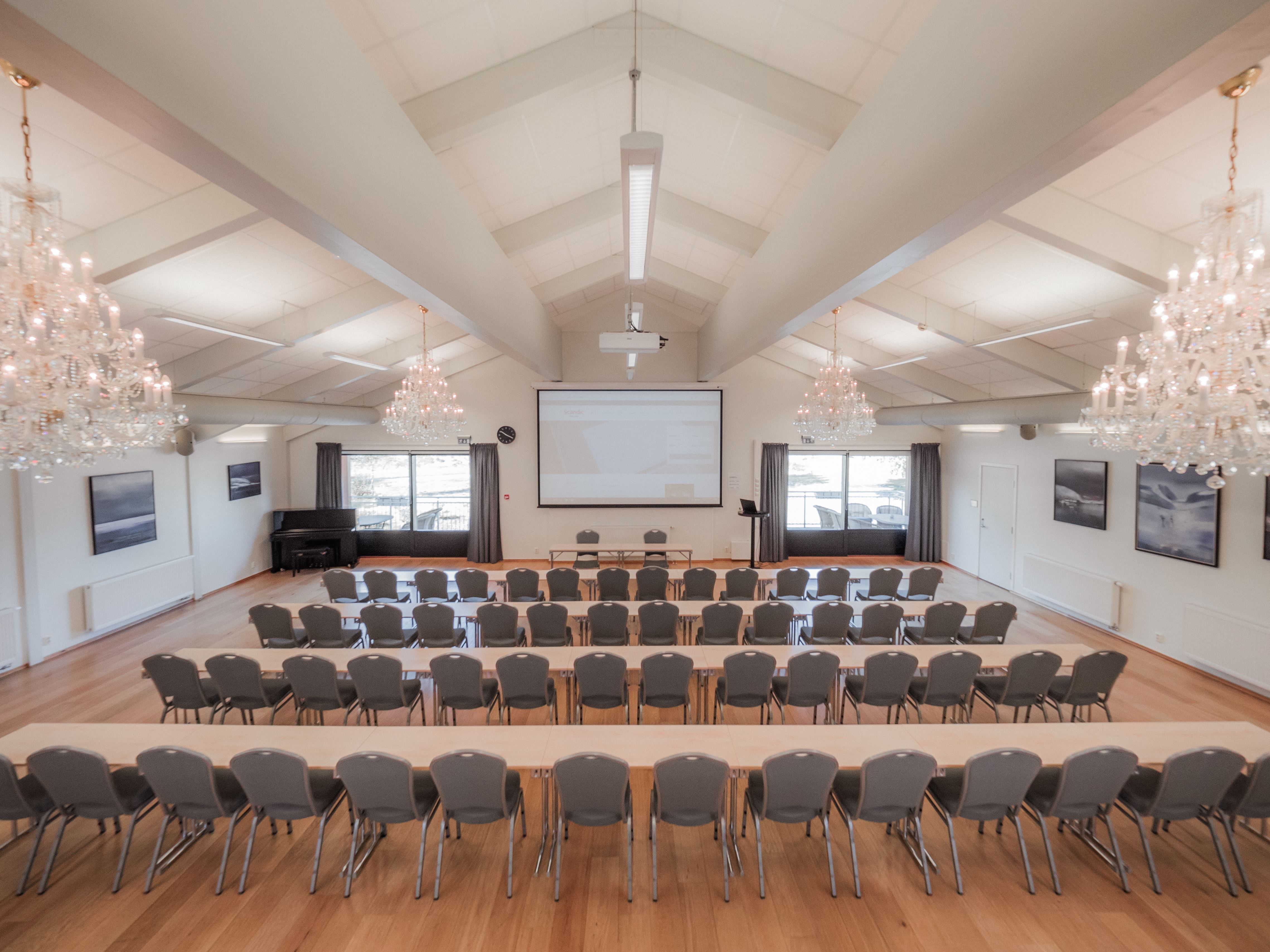 Bright conference room with rows of tables and chairs facing a projector screen, chandeliers, and large windows.