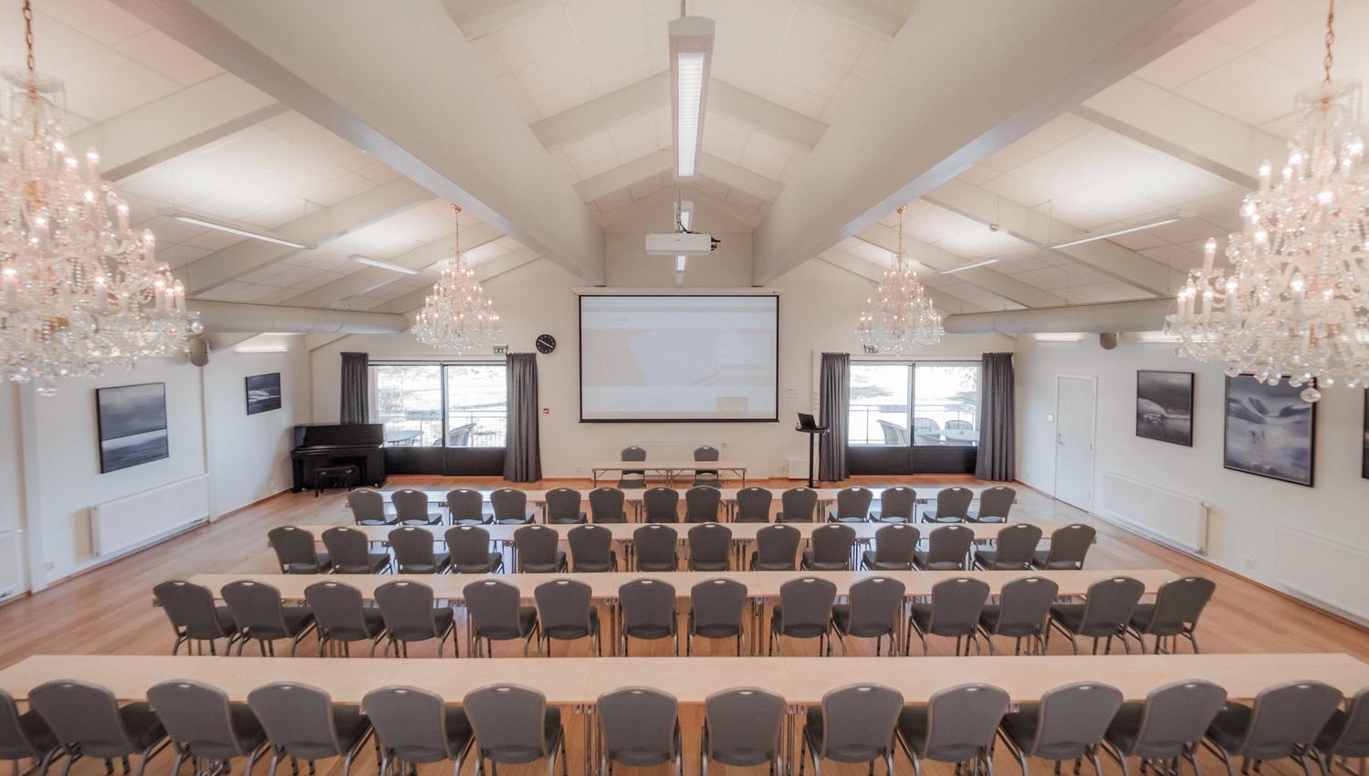 Bright conference room with rows of tables and chairs facing a projector screen, chandeliers, and large windows.