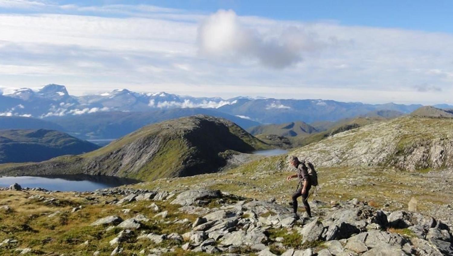 A lone hiker with a backpack walks across a rocky ridge above small lakes, with distant mountains and clouds.