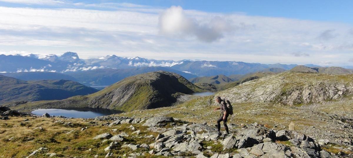 A lone hiker with a backpack walks across a rocky ridge above small lakes, with distant mountains and clouds.