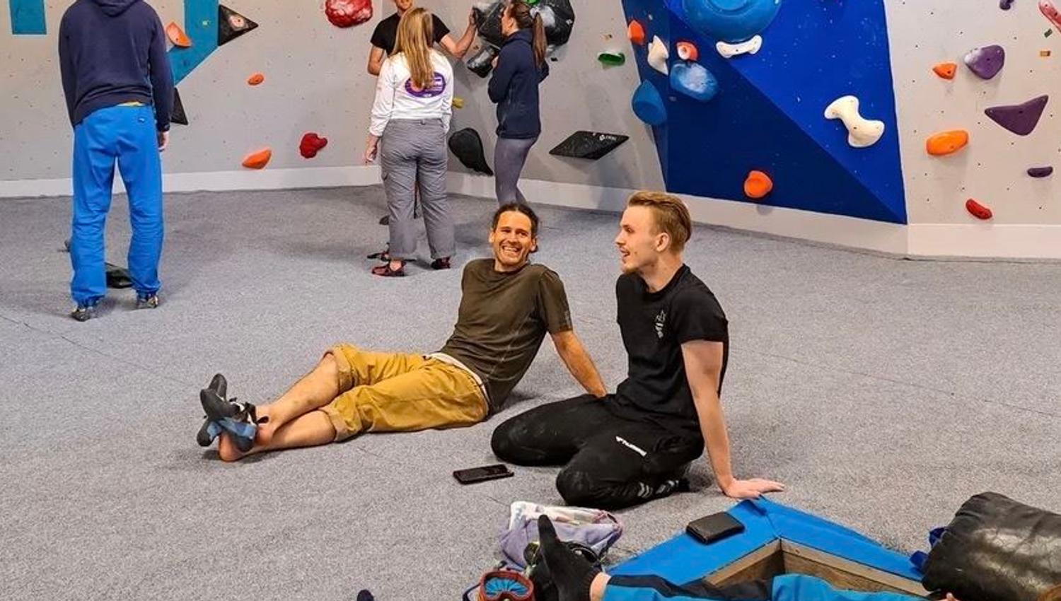People rest and chat on padded flooring inside an indoor bouldering gym with colorful climbing holds on walls..