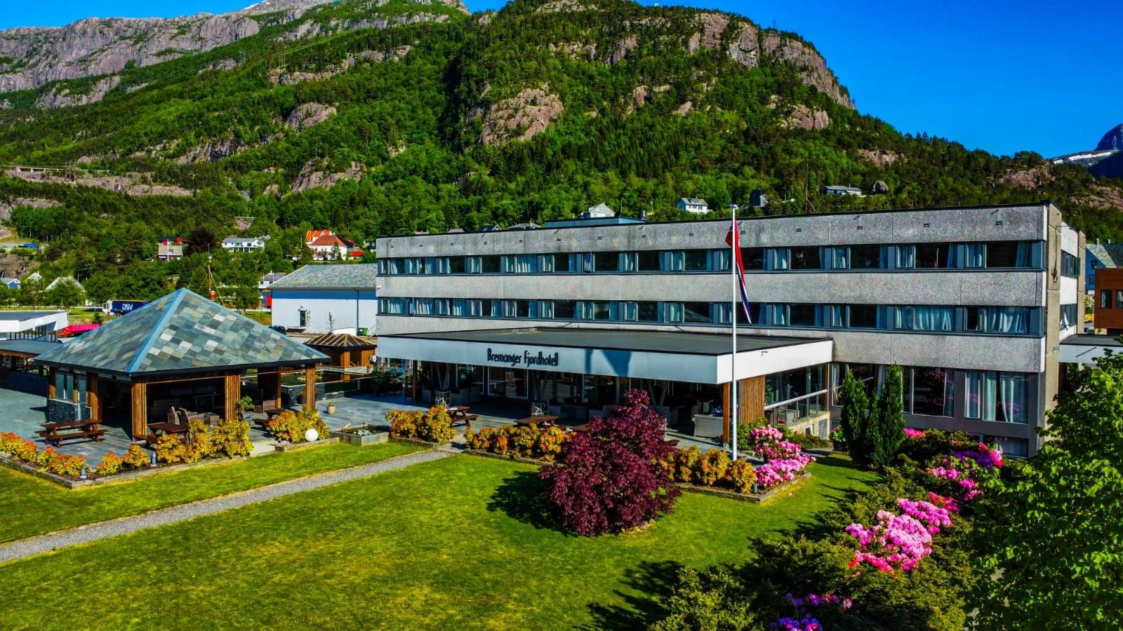 Stort hotellbygg med uteplass, blomsterbed og flaggstong, med grøne fjell og blå himmel i bakgrunnen..