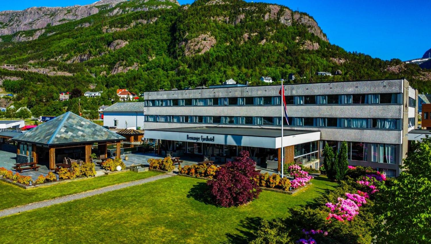 Stort hotellbygg med uteplass, blomsterbed og flaggstong, med grøne fjell og blå himmel i bakgrunnen..