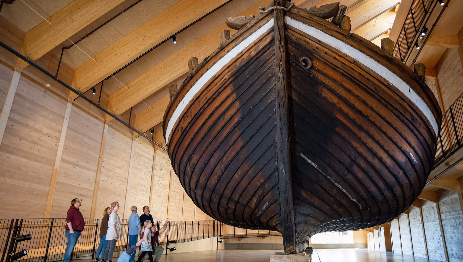 Large wooden boat hull displayed indoors on supports, with visitors standing and walking beneath high timber beams..