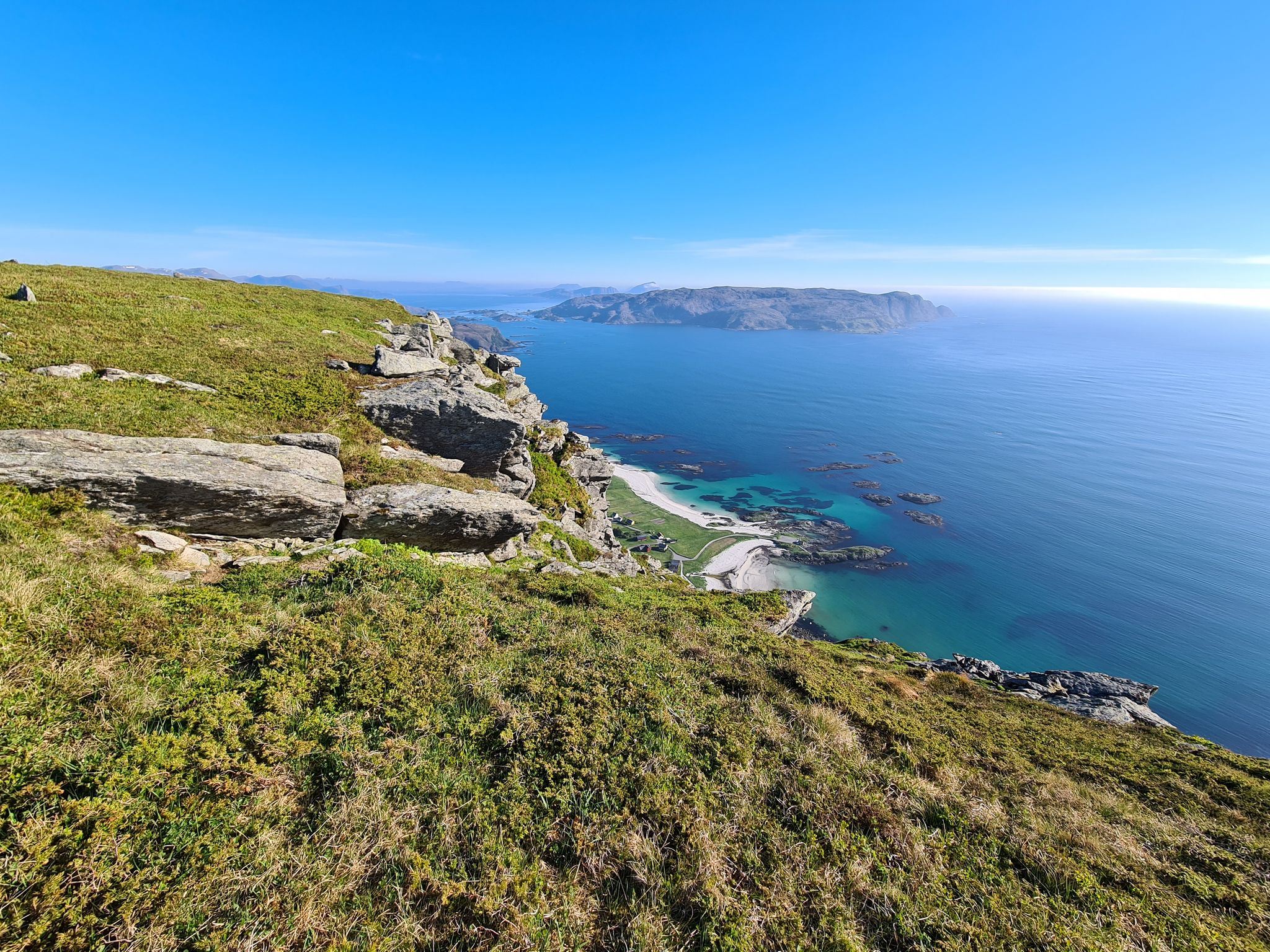Grassy rocky cliff overlooks a turquoise bay with small islands and a distant headland under a clear blue sky.