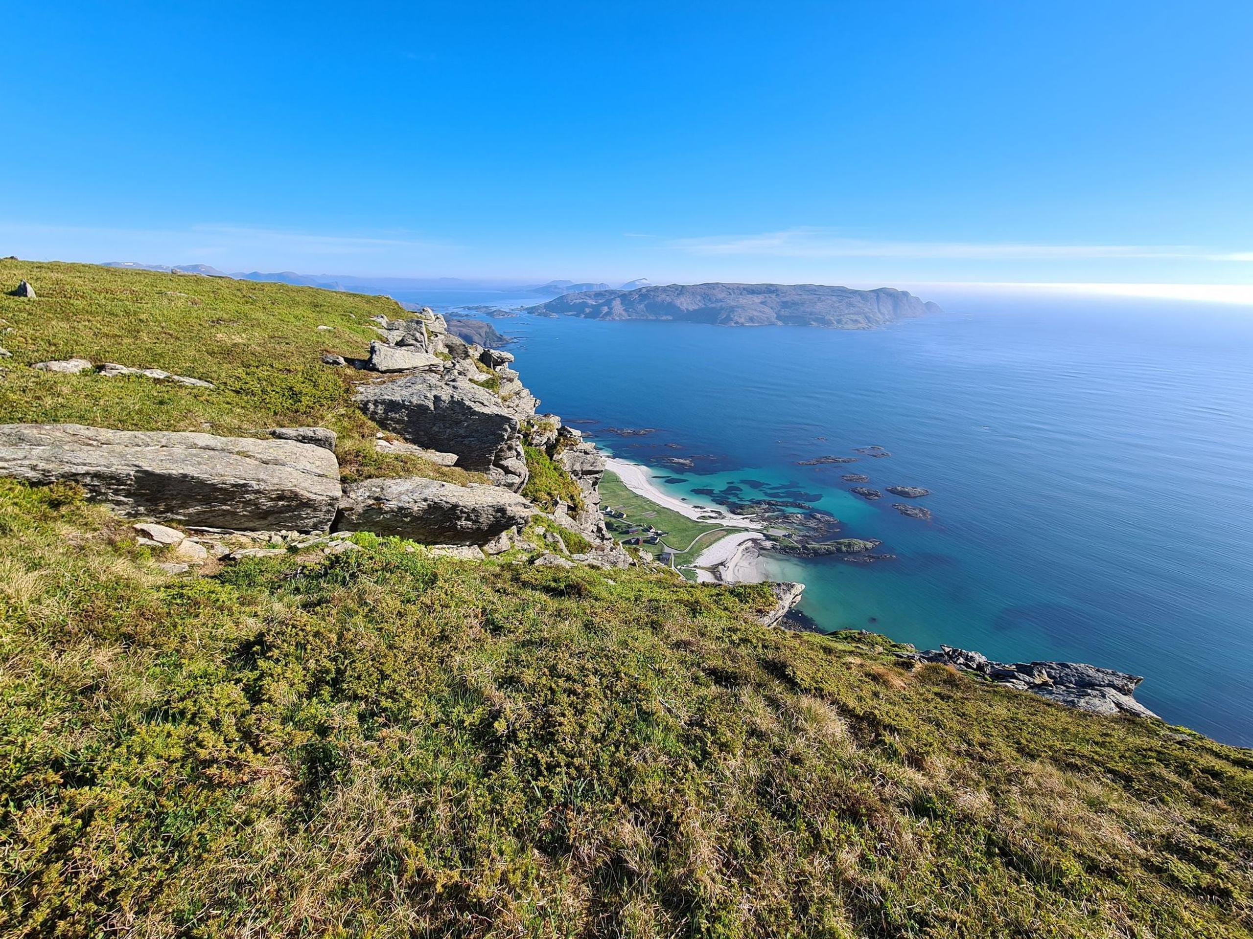 Grassy rocky cliff overlooks a turquoise bay with small islands and a distant headland under a clear blue sky.