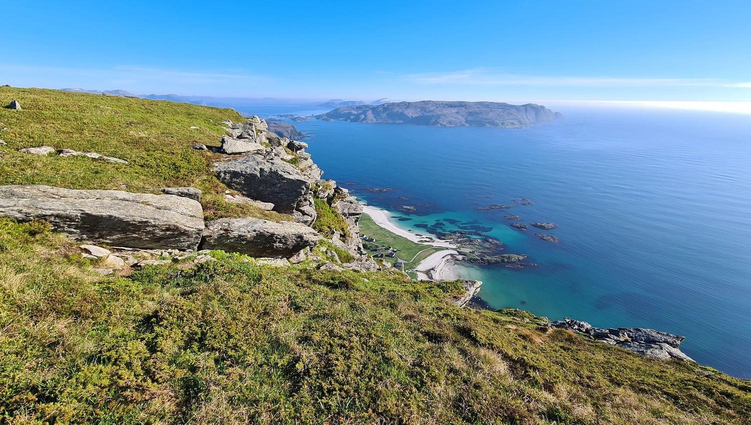 Grassy rocky cliff overlooks a turquoise bay with small islands and a distant headland under a clear blue sky.