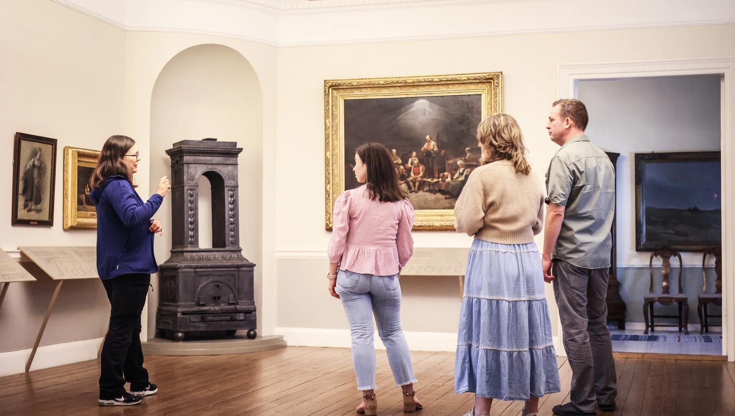 Four people are inside Mandal Museum looking at the large painting “The Haugeans”, where Adolph Tidemand depicts the followers of the lay preacher Han