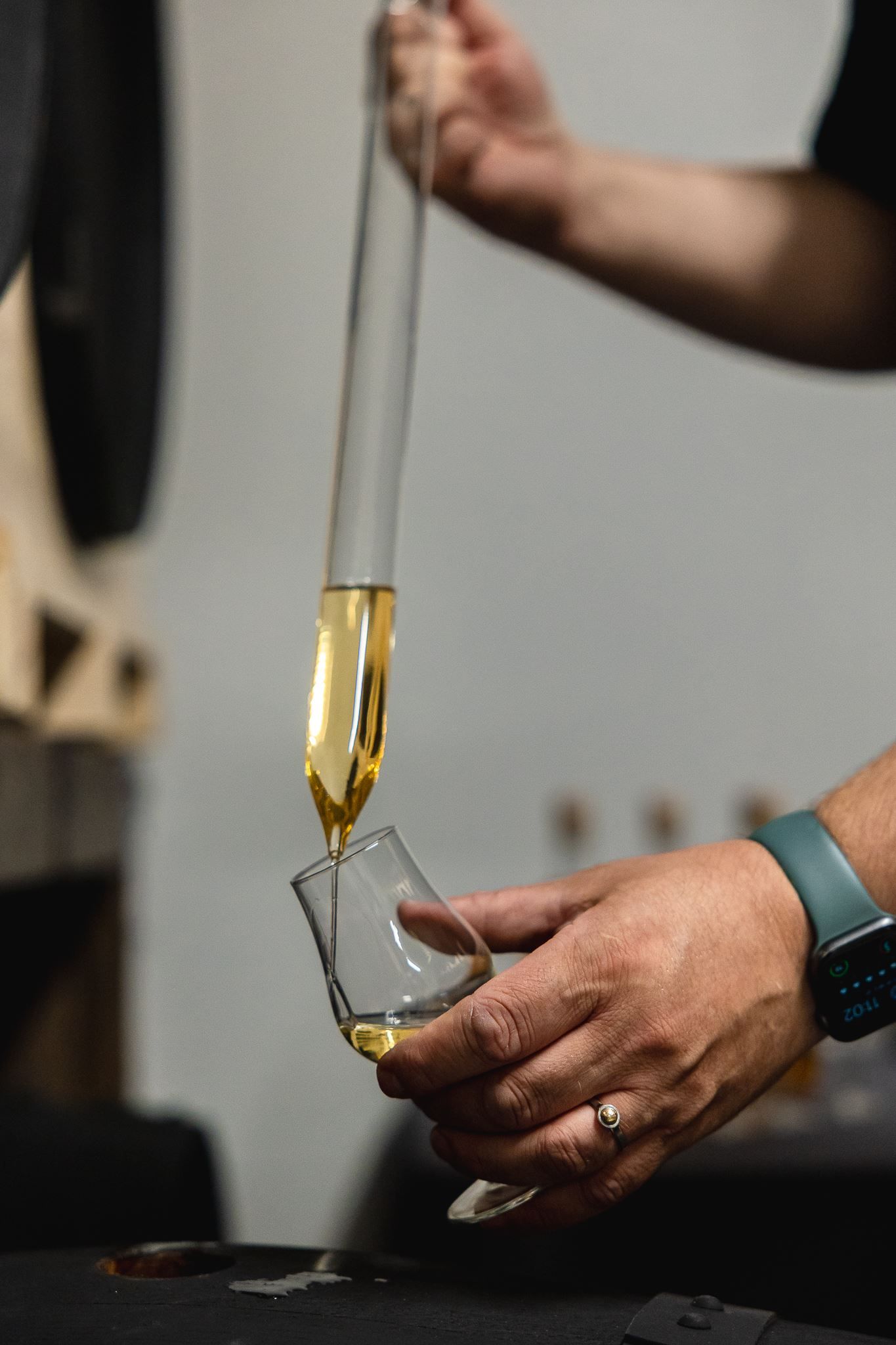 Close-up of hands using a glass pipette to pour amber liquor into a tilted tasting glass indoors.