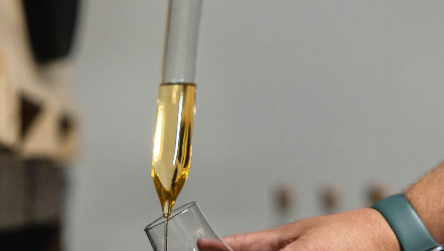 Close-up of hands using a glass pipette to pour amber liquor into a tilted tasting glass indoors.