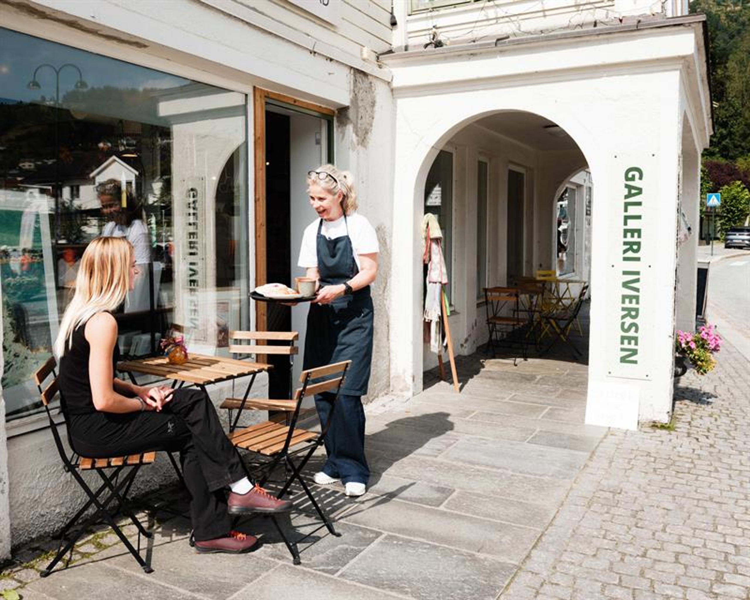 A woman is seated at a small outdoor café table while being served coffee and baked goods outside Iversen Bakery and Gallery in Norheimsund, with the 