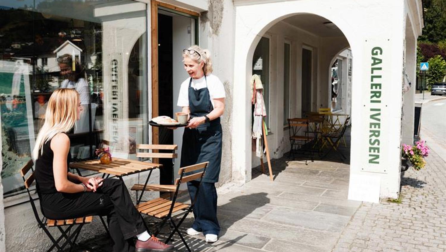 A woman is seated at a small outdoor café table while being served coffee and baked goods outside Iversen Bakery and Gallery in Norheimsund, with the