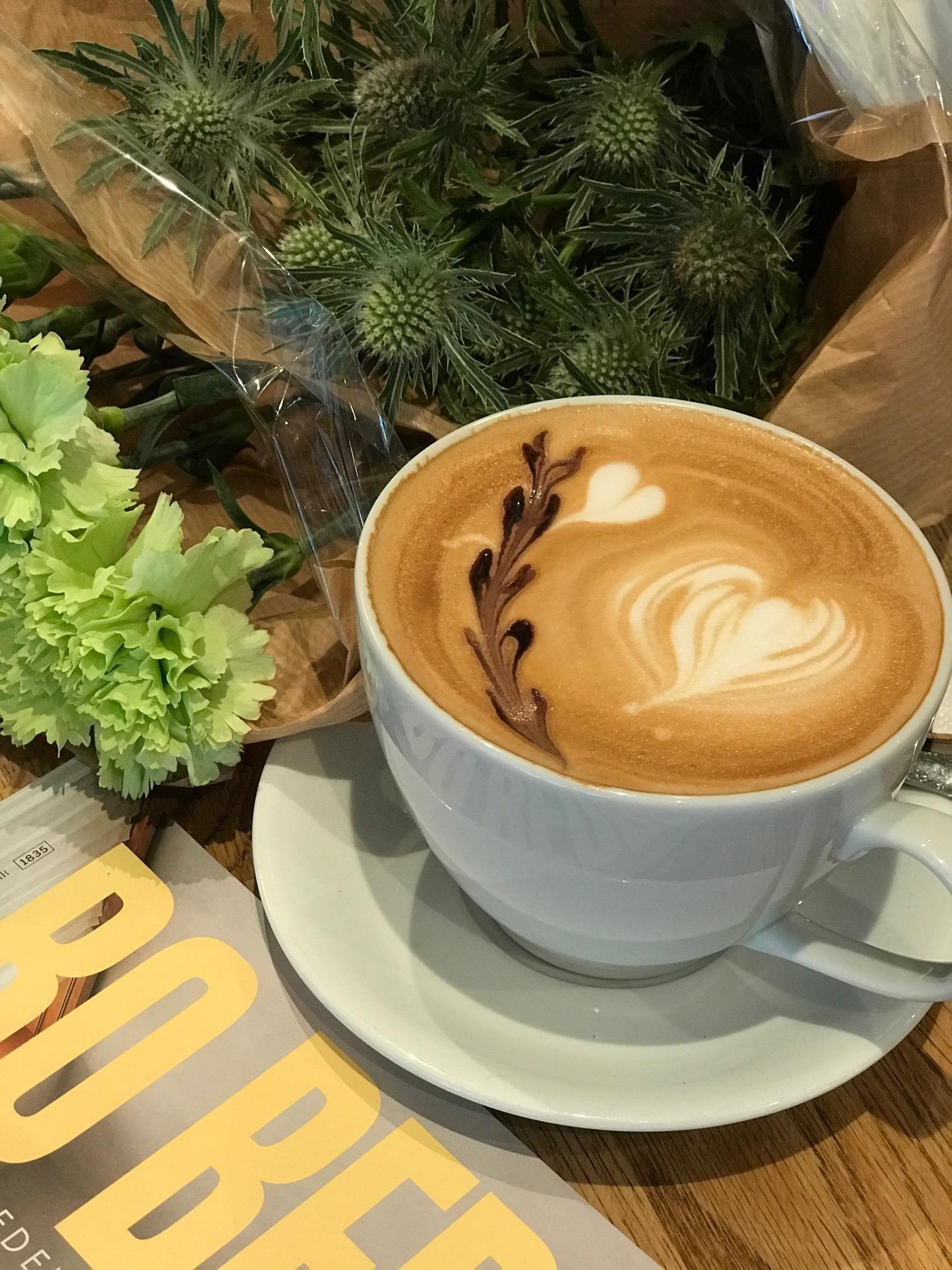 White cup of latte with heart latte art on a saucer beside a bouquet of green flowers on a wooden table.