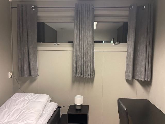 Compact bedroom with a bed, bedside table and lamp, and a high window framed by gray curtains.