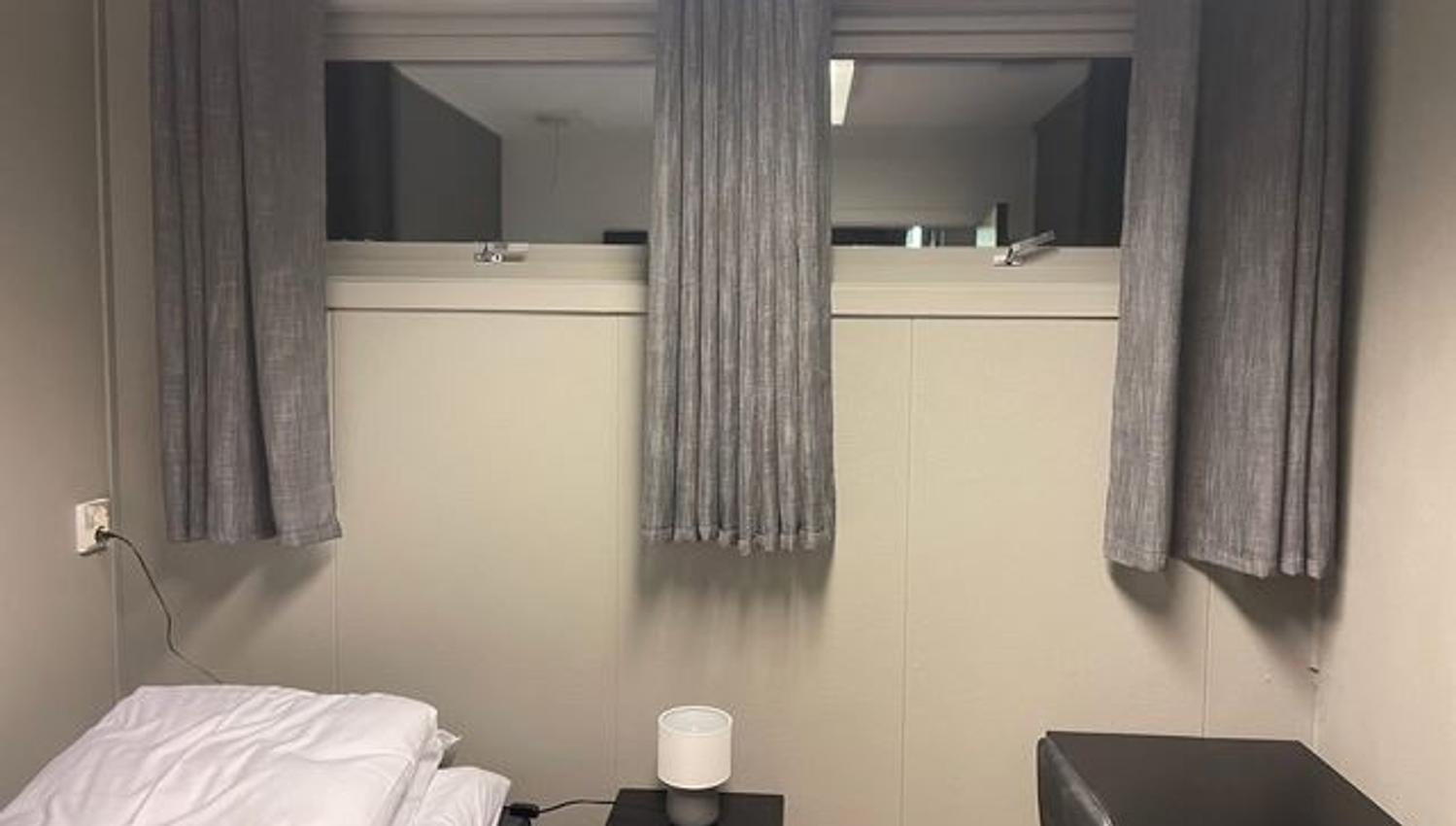Compact bedroom with a bed, bedside table and lamp, and a high window framed by gray curtains.