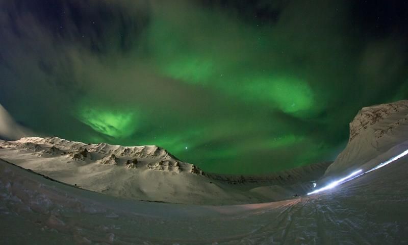 Grønt nordlys fyller himmelen over fjellene i snødekt landskap under polarnatten