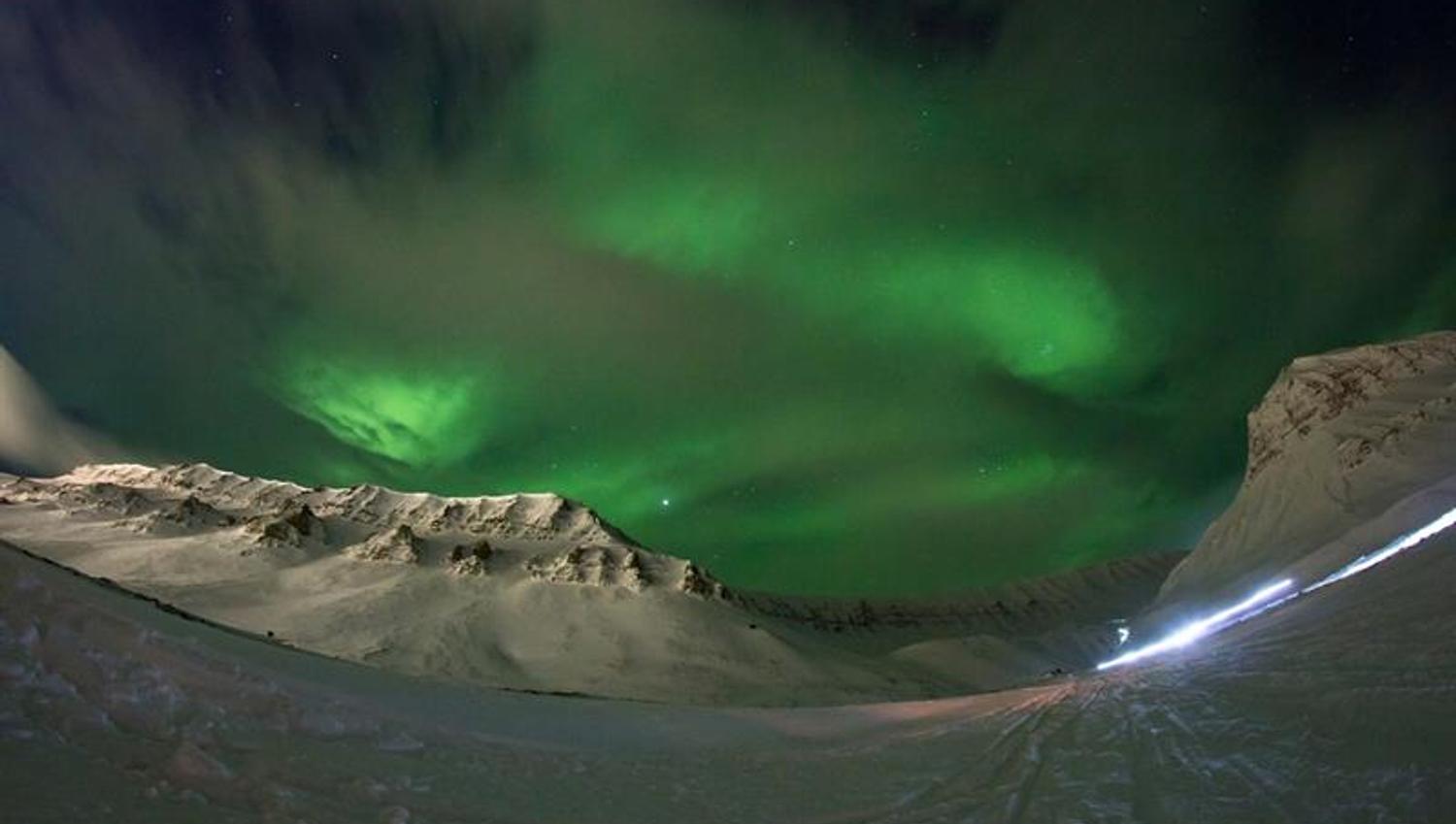 Grønt nordlys fyller himmelen over fjellene i snødekt landskap under polarnatten