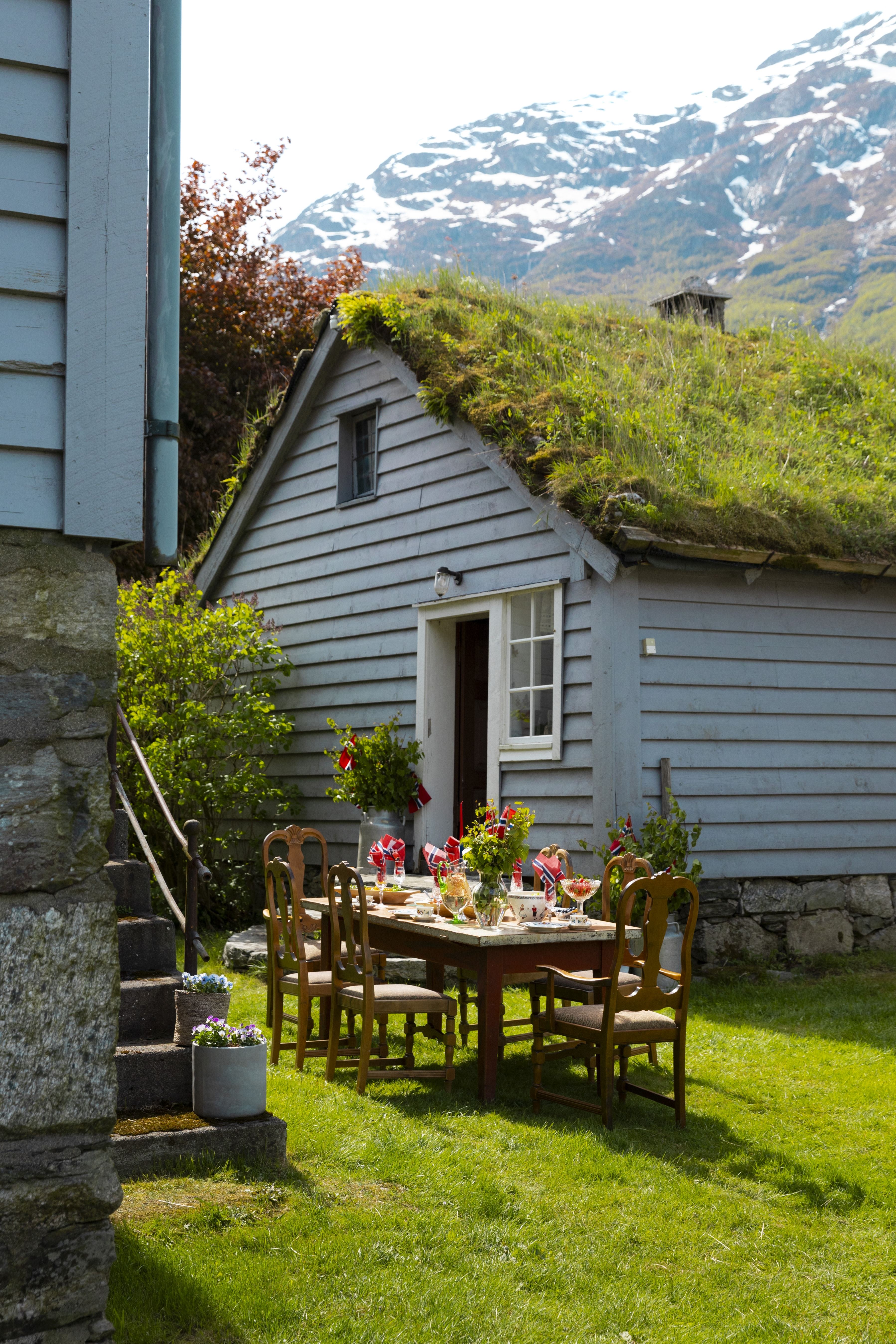 Outdoor dining table with wooden chairs on a green lawn beside a small cottage with a grass roof and snowy mountains behind.