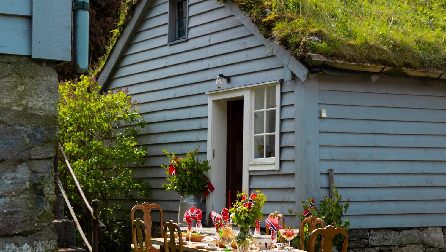 Outdoor dining table with wooden chairs on a green lawn beside a small cottage with a grass roof and snowy mountains behind.