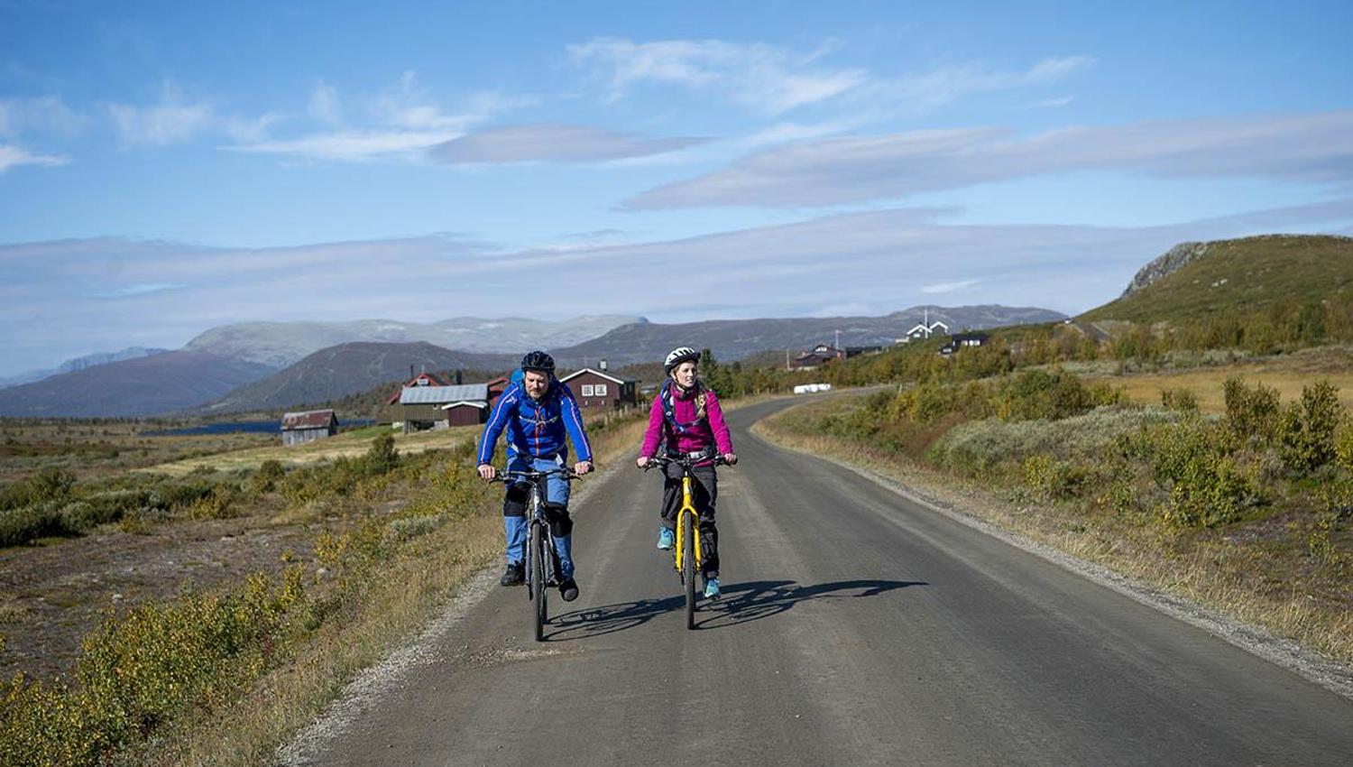 To syklister i fargerike jakker på sykkeltur på Mjølkevegen over Stølsvidda, i fritt og høytliggende landskap.