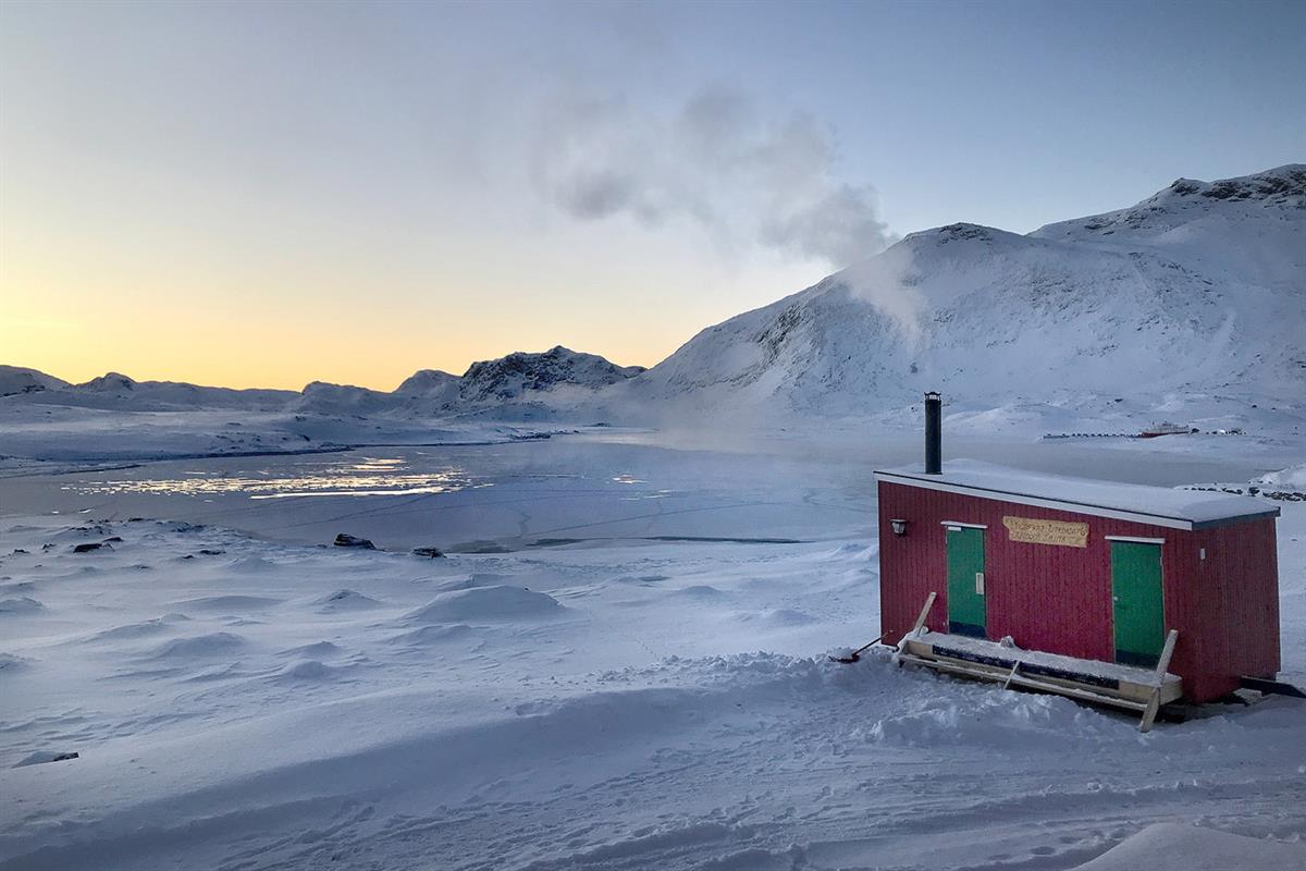 Small red outdoor sauna on a lakeshore in winter with a snowy mountain landscape