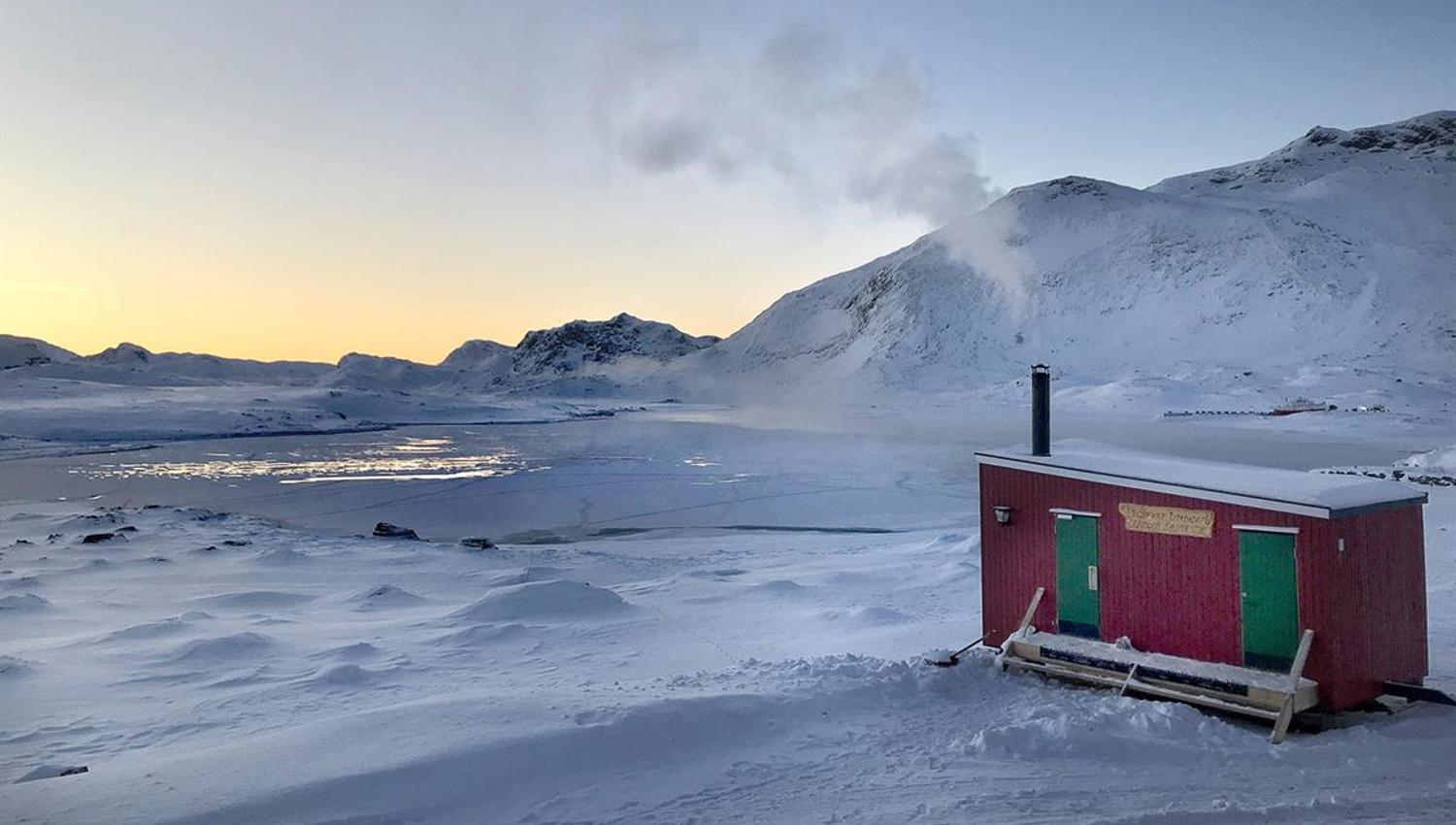 Small red outdoor sauna on a lakeshore in winter with a snowy mountain landscape