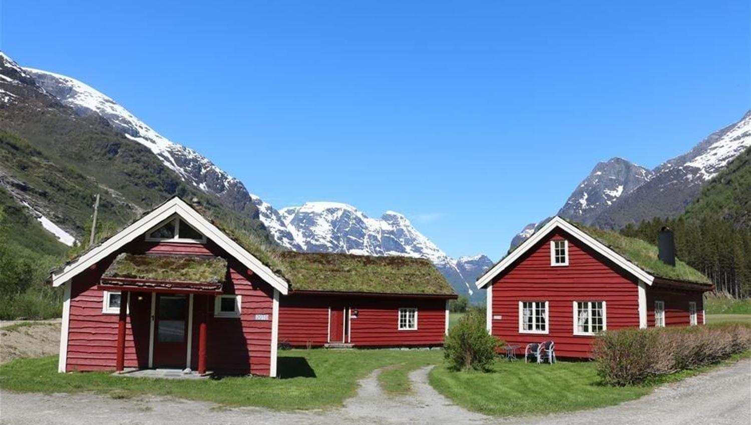 Two red wooden cabins with grass roofs sit on green grass, framed by snowy mountains under a clear blue sky at Aabrekk Gard Trollbu in Oldedalen valley..