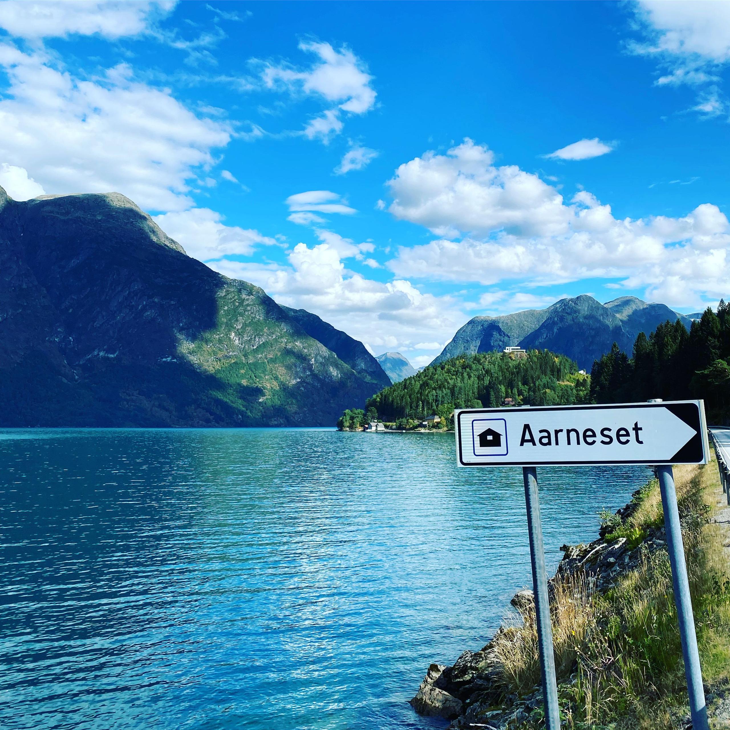 Vegskilt med teksten «Aarneset» ved ein blå fjord, med fjell og skyer i bakgrunnen på ein klår dag..