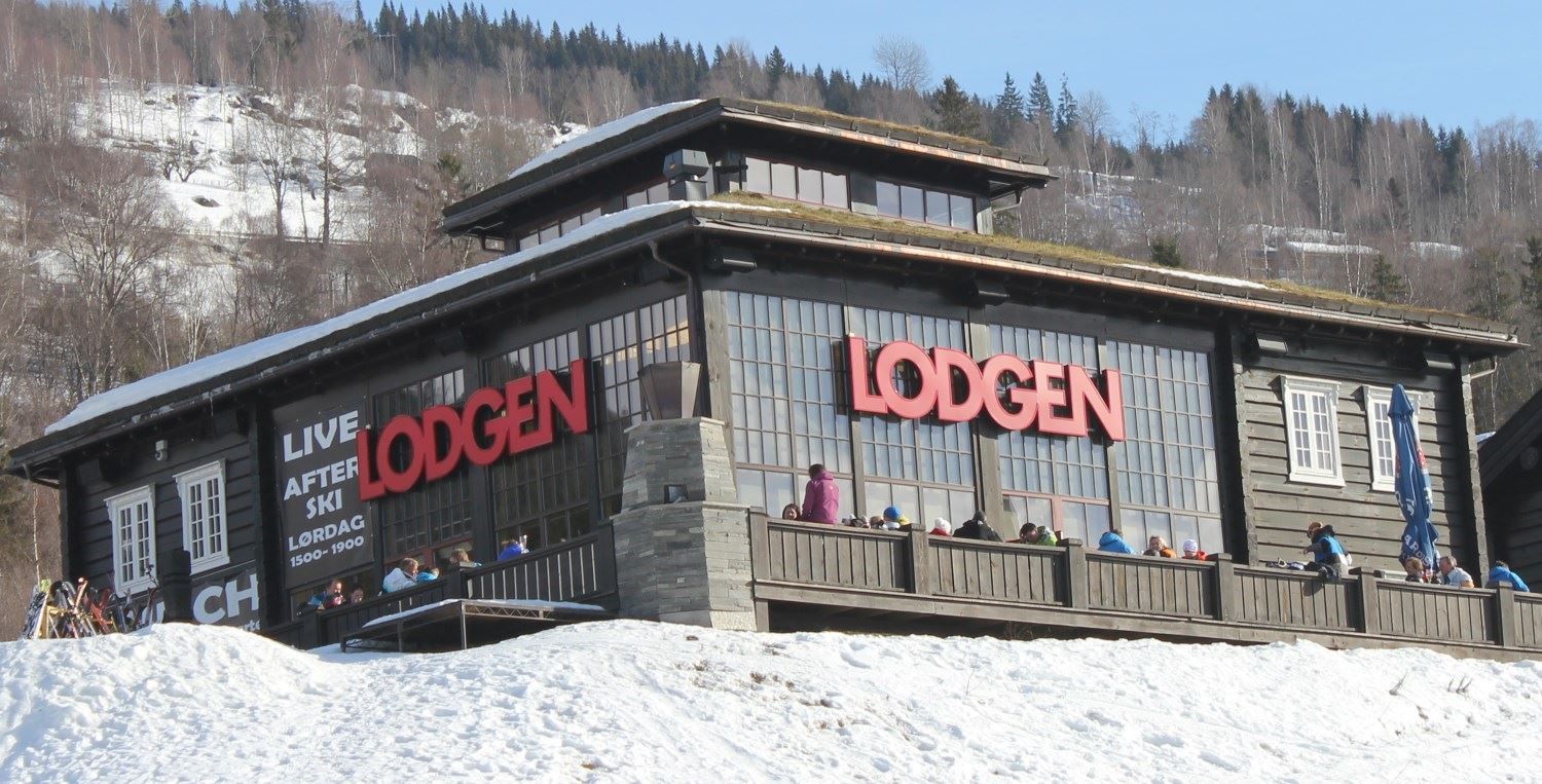 Moderne trehytte med store vinduer og rødt LODGEN-skilt, folk på terrasse, snø i forgrunn og skogkledd fjell bak..