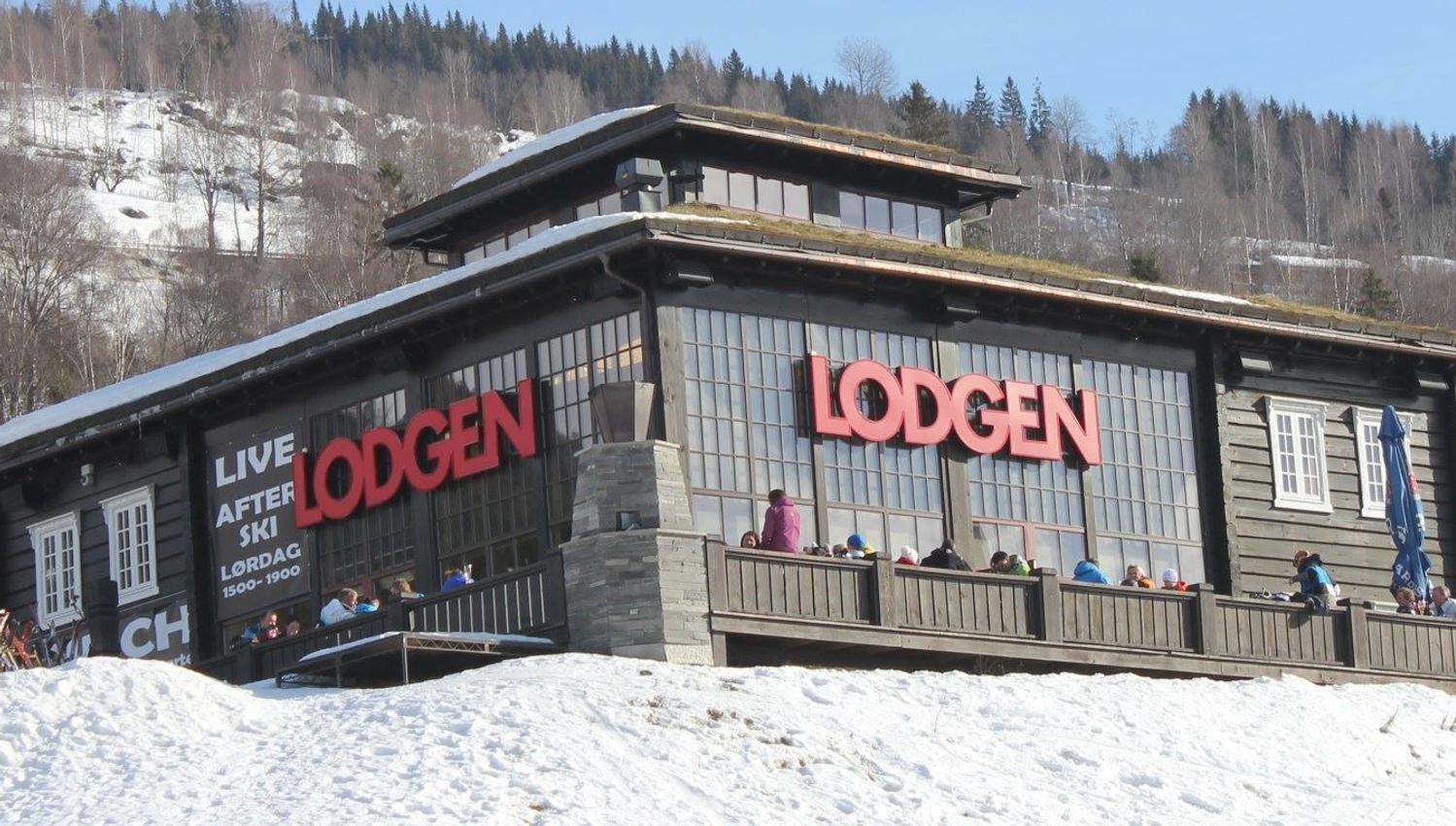 Moderne trehytte med store vinduer og rødt LODGEN-skilt, folk på terrasse, snø i forgrunn og skogkledd fjell bak..