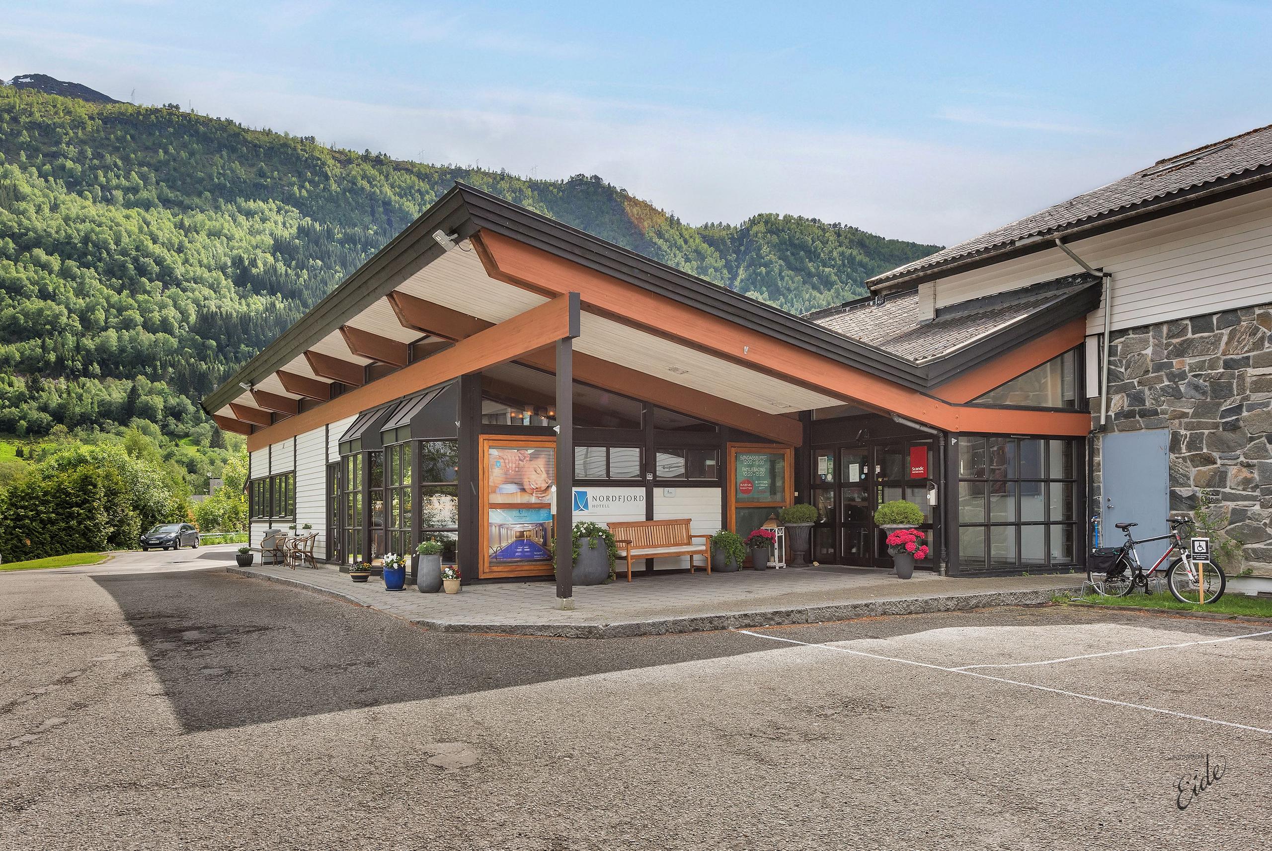 Modern lodge entrance with wooden beams, glass doors, potted plants, and forested mountains under a clear sky.