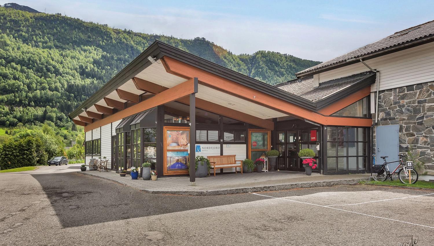 Modern lodge entrance with wooden beams, glass doors, potted plants, and forested mountains under a clear sky.
