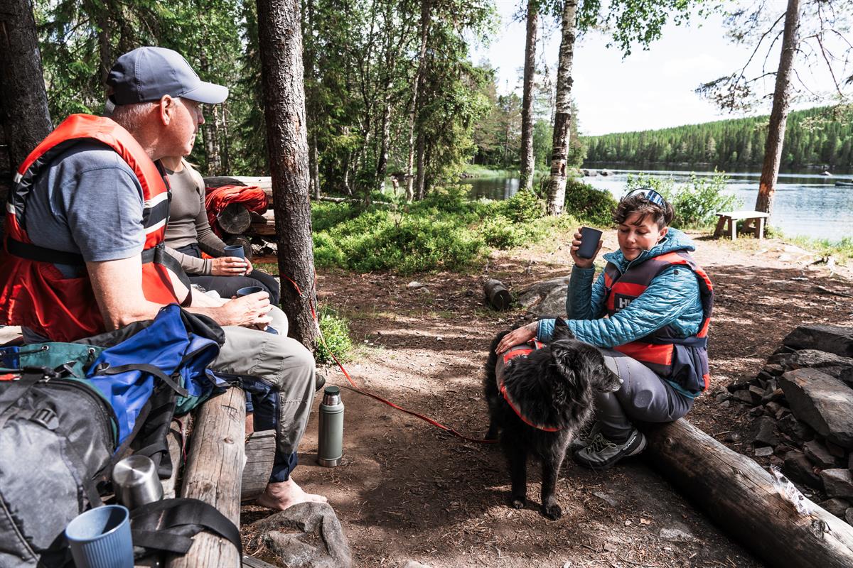 Three people resting at a forest campsite by the water, one petting a black dog wearing a life vest.