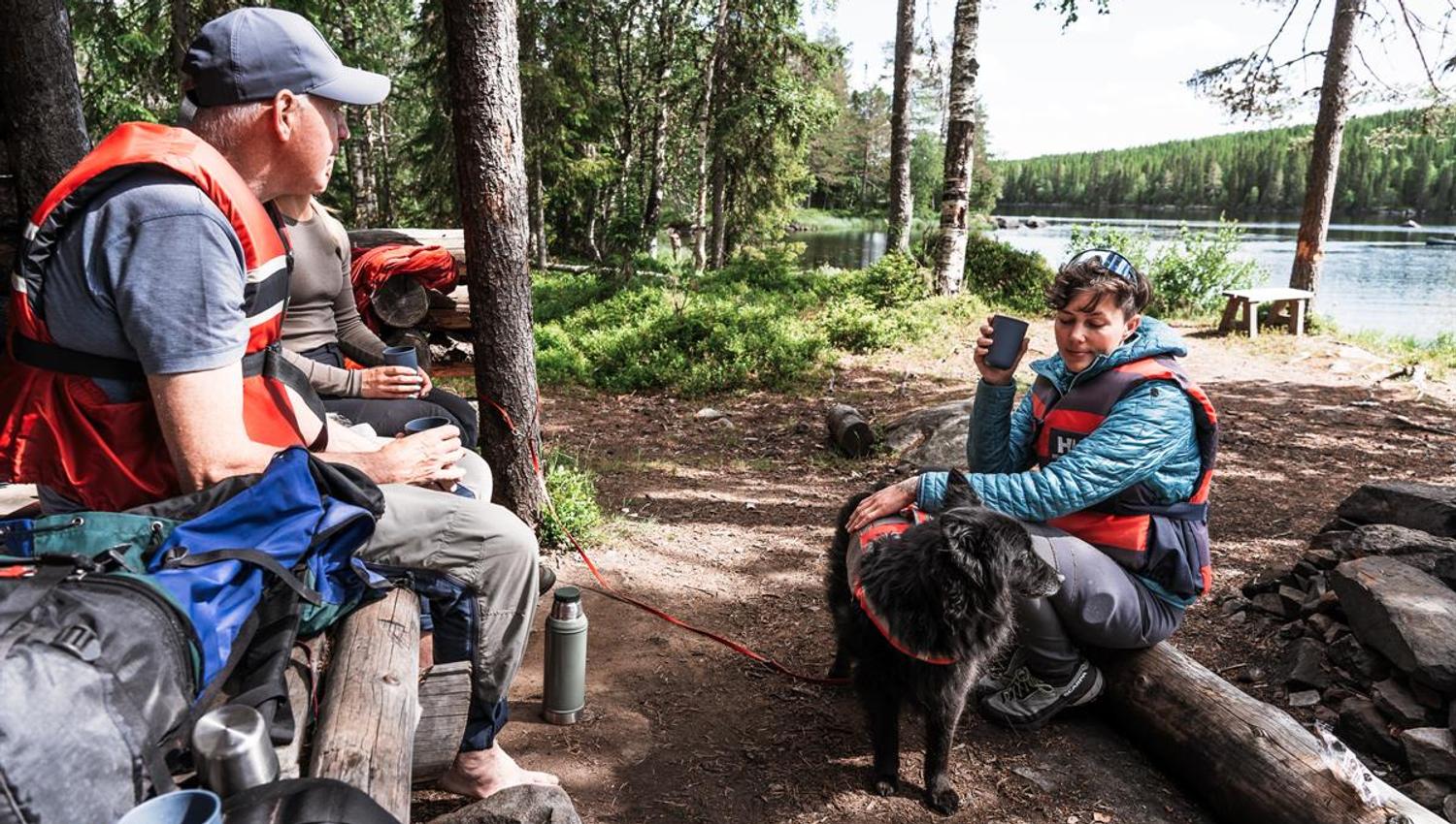 Three people resting at a forest campsite by the water, one petting a black dog wearing a life vest.