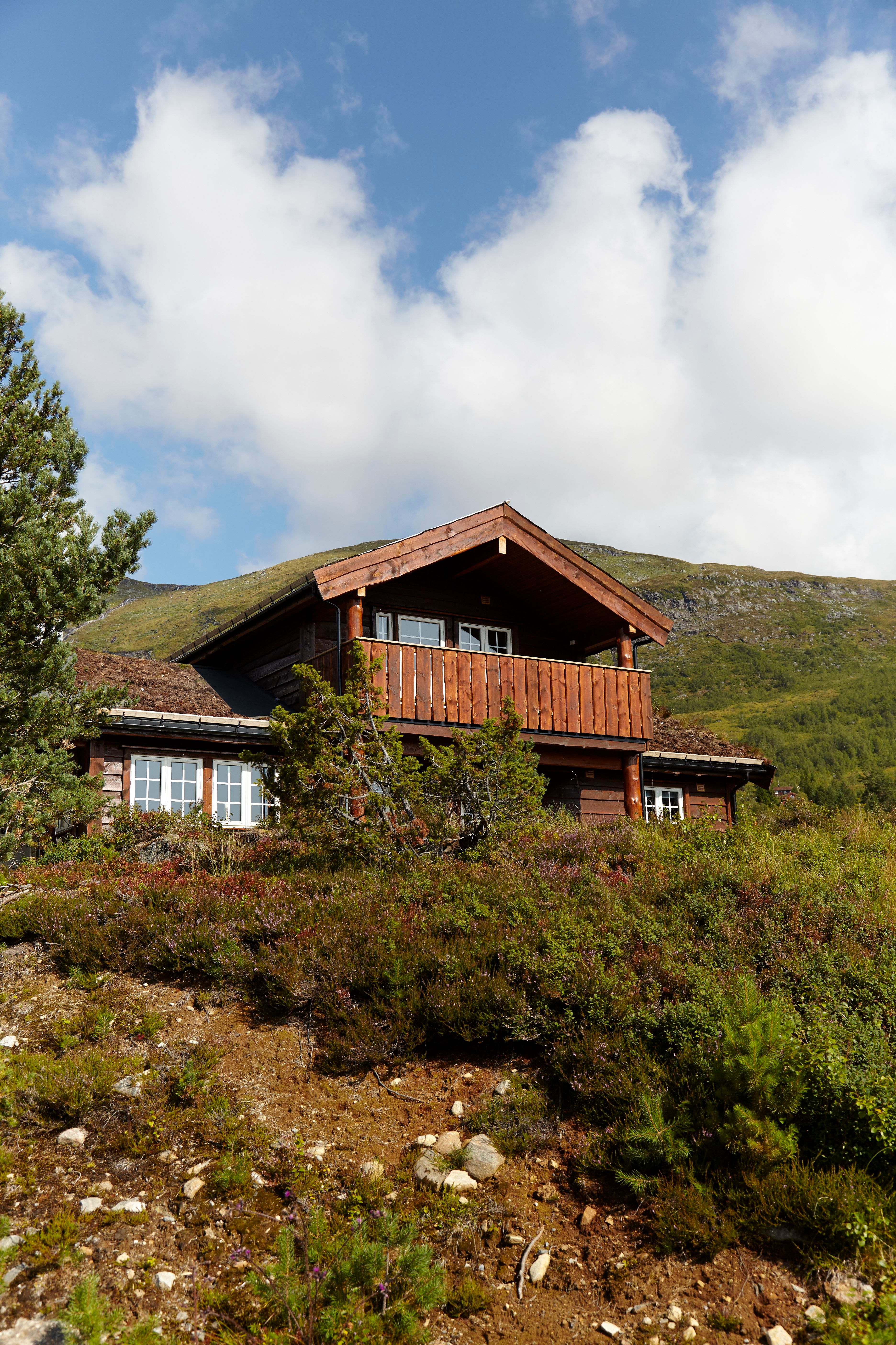 Furuvins Lodge med balkong ved Stryn sine turløyper i fjellterreng, omgitt av lyng og busker under blå himmel med skyer..