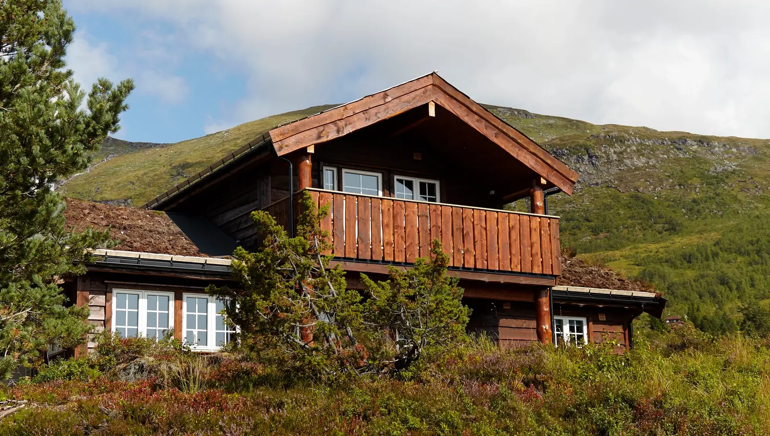 Furuvins Lodge med balkong ved Stryn sine turløyper i fjellterreng, omgitt av lyng og busker under blå himmel med skyer..