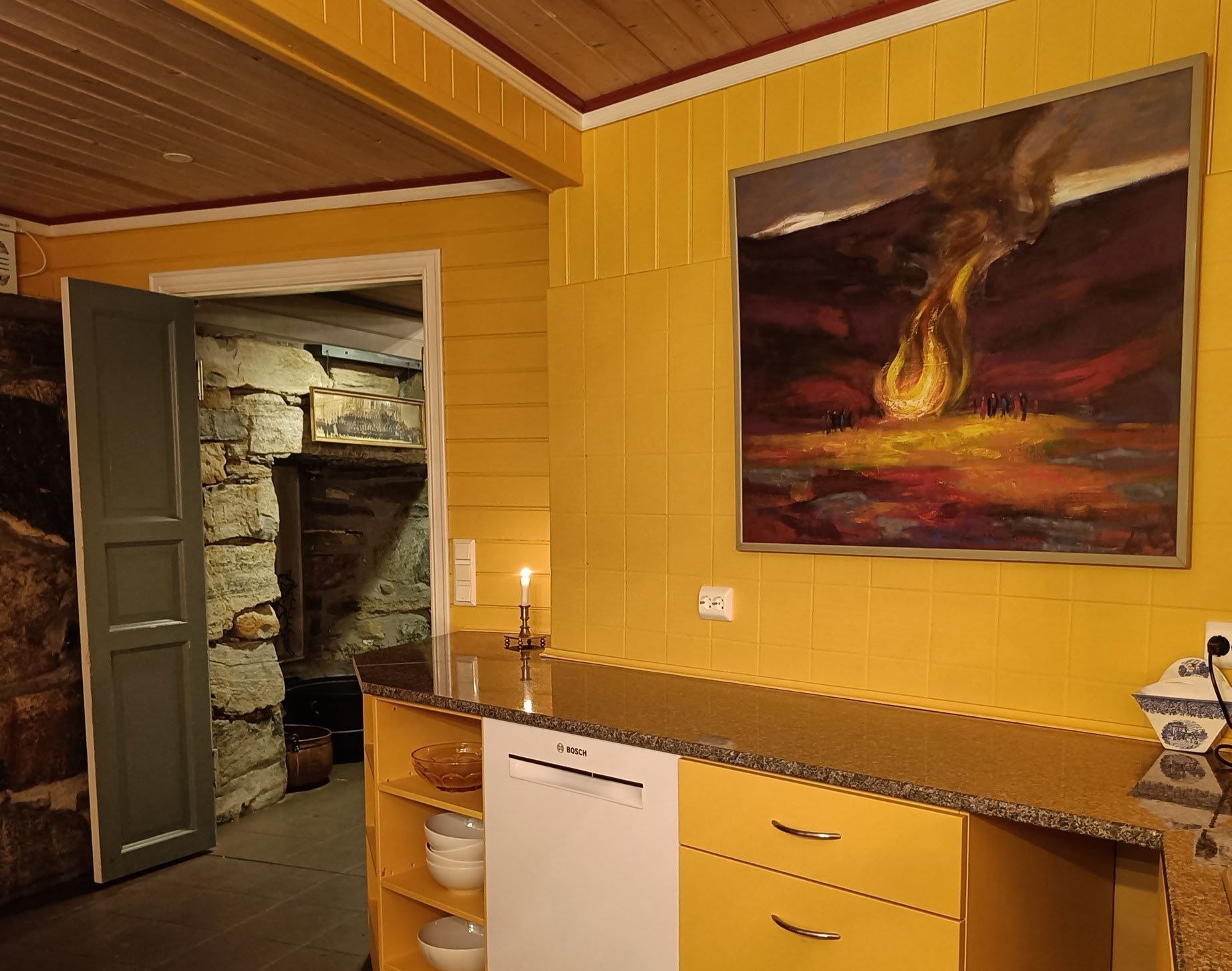 Orheimstunet in Nordfjord - Yellow-paneled kitchen with countertop, dishwasher, candle, and large painting; open door reveals stone room beyond..