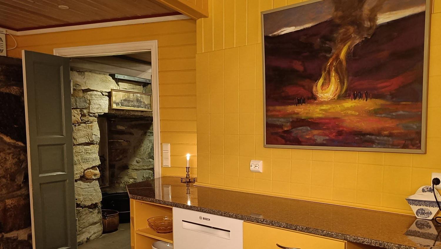 Orheimstunet in Nordfjord - Yellow-paneled kitchen with countertop, dishwasher, candle, and large painting; open door reveals stone room beyond..
