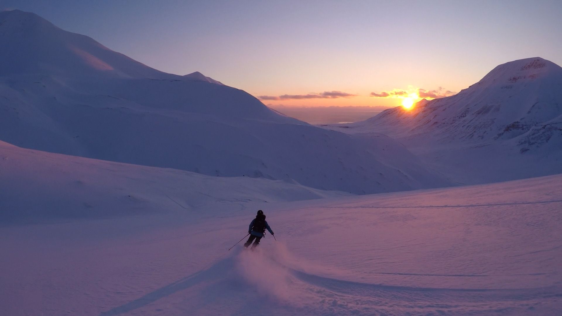 A person skiing down a mountain at dusk with a sunset over a mountain ridge in the background